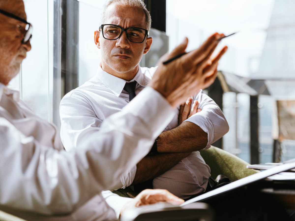 Two male executives are engaged in a focused conversation in a modern office setting. Both are wearing glasses and white dress shirts, and one man is gesturing with a pen while explaining something, while the other listens attentively with his arms crossed.