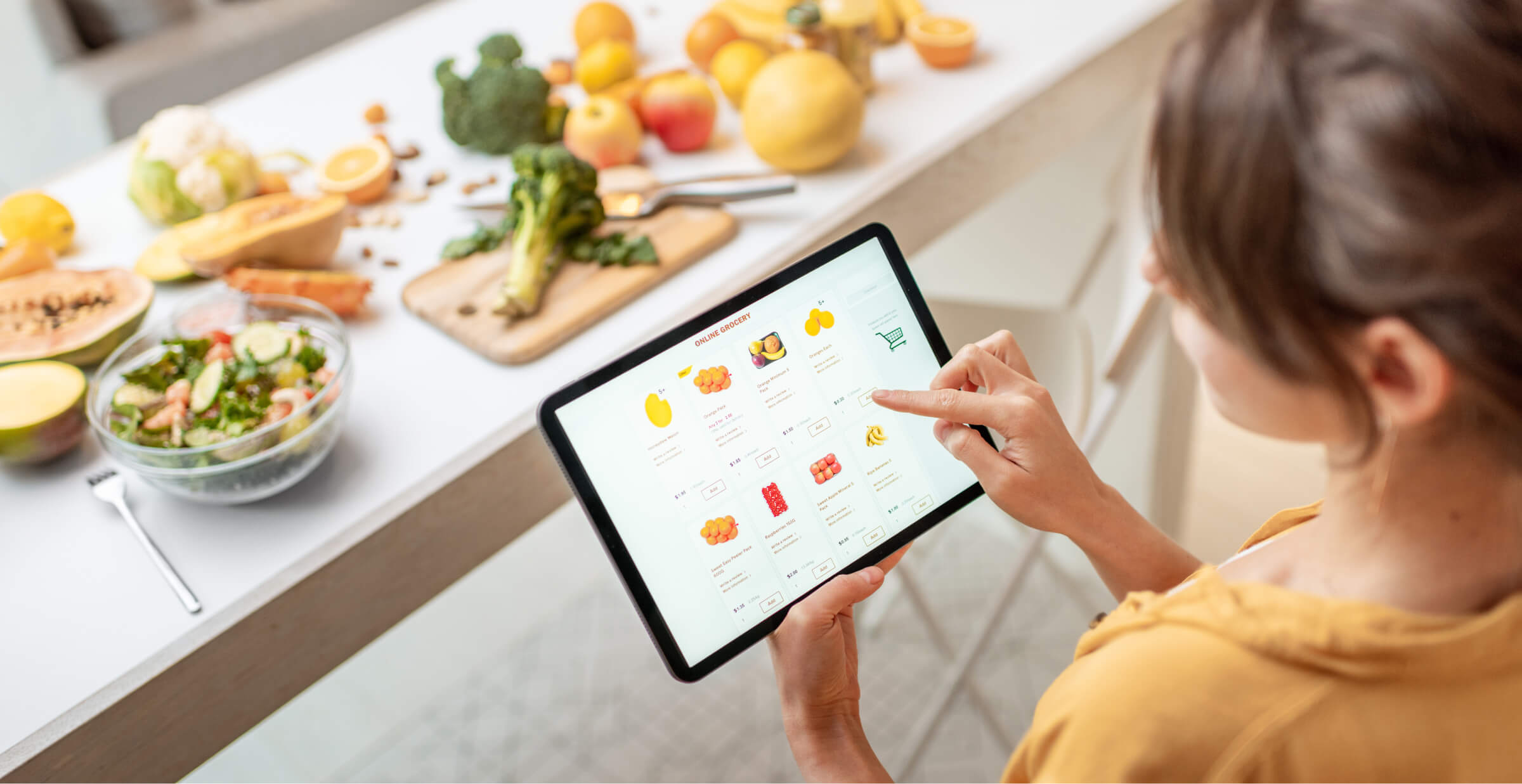 A woman using a tablet to shop for groceries online while standing in a kitchen. The kitchen counter is filled with fresh fruits, vegetables, and a salad, creating a healthy and vibrant atmosphere. The tablet screen displays various grocery items, emphasizing the convenience of online grocery shopping.