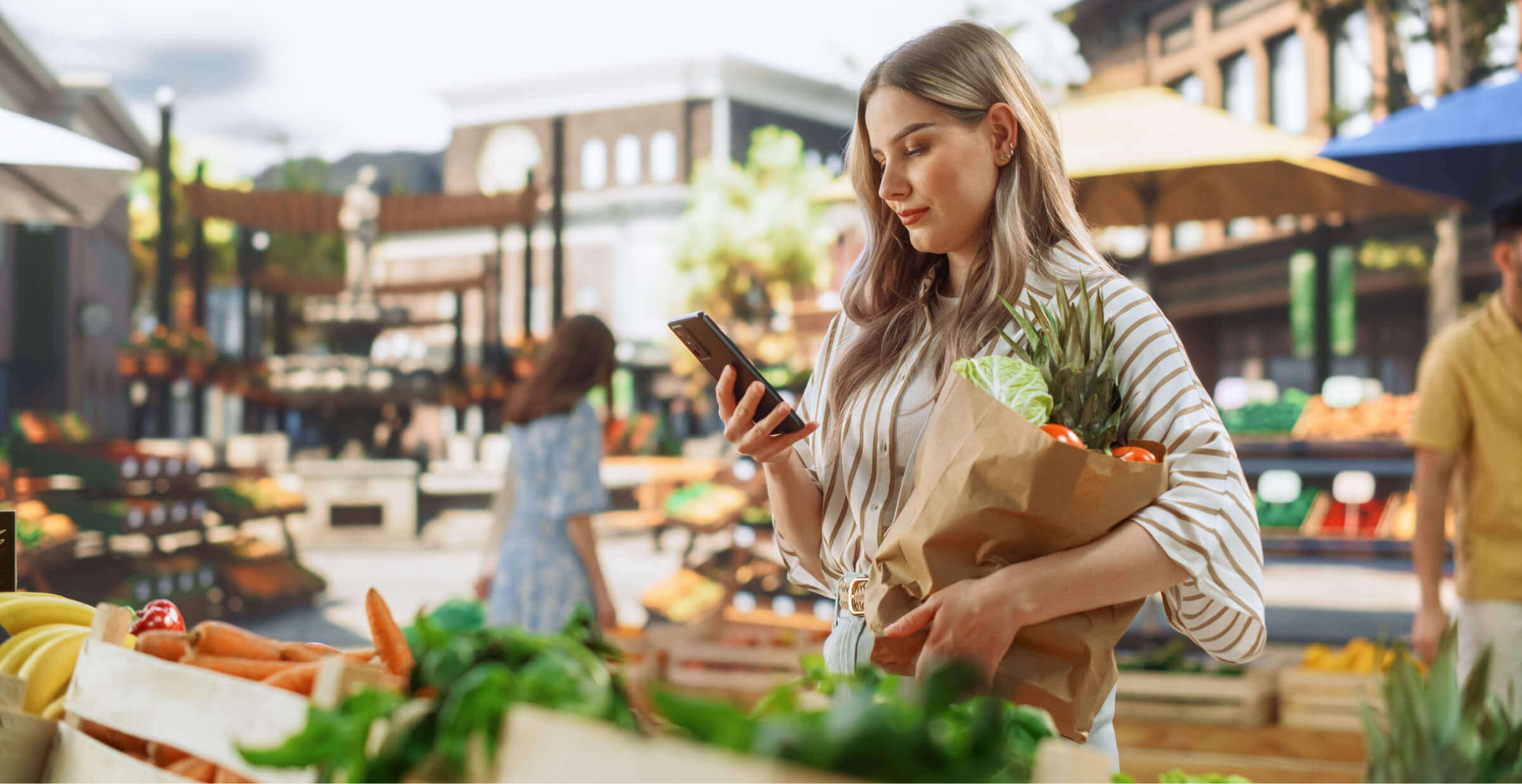 A woman shopping at an outdoor market, holding a paper bag filled with fresh produce such as pineapples and tomatoes. She is looking at her smartphone, possibly using it for a shopping list or payment, surrounded by vibrant market stalls filled with fruits and vegetables.