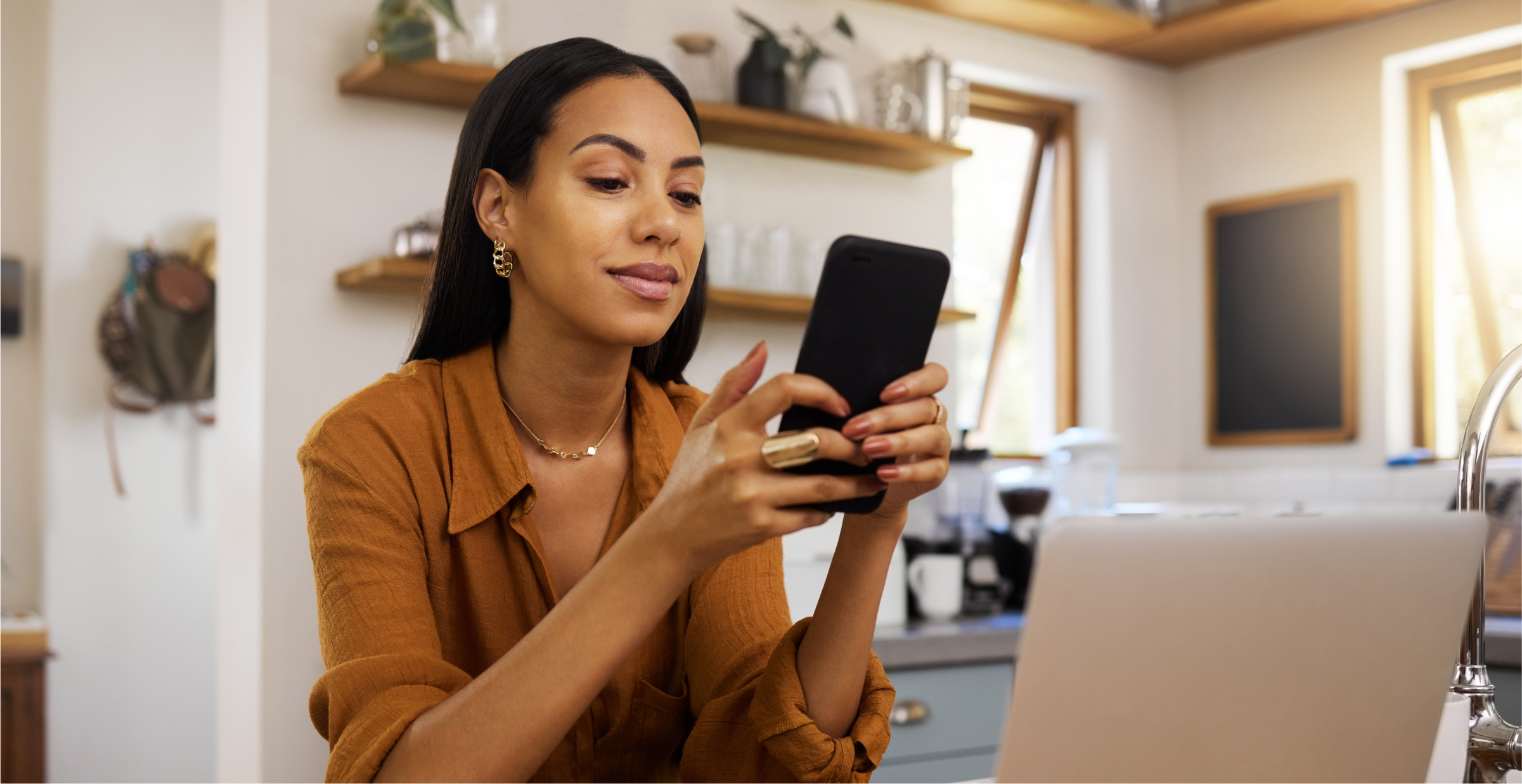 A woman is sitting in a cozy home office, smiling as she uses her smartphone. She appears relaxed and focused, with a laptop in front of her and a modern kitchen in the background. The setting suggests a comfortable and productive work-from-home environment, with natural light streaming through the windows. The image highlights the balance of professional and personal life in a home office setting.