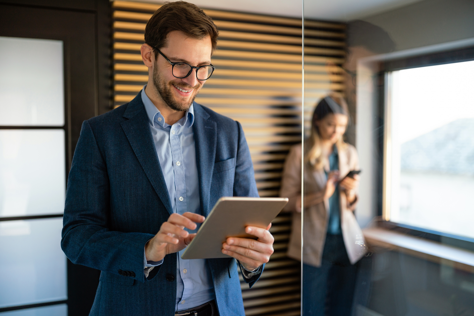 A man in a business suit and glasses is smiling while working on a tablet in a modern office. He is standing near a glass wall, with a woman in the background using her phone. The man appears focused and engaged, likely reviewing or analyzing something on the tablet. The setting is professional, with warm lighting and a sleek design, featuring wooden panels on the wall. The image conveys a sense of productivity, technology use, and a collaborative work environment.