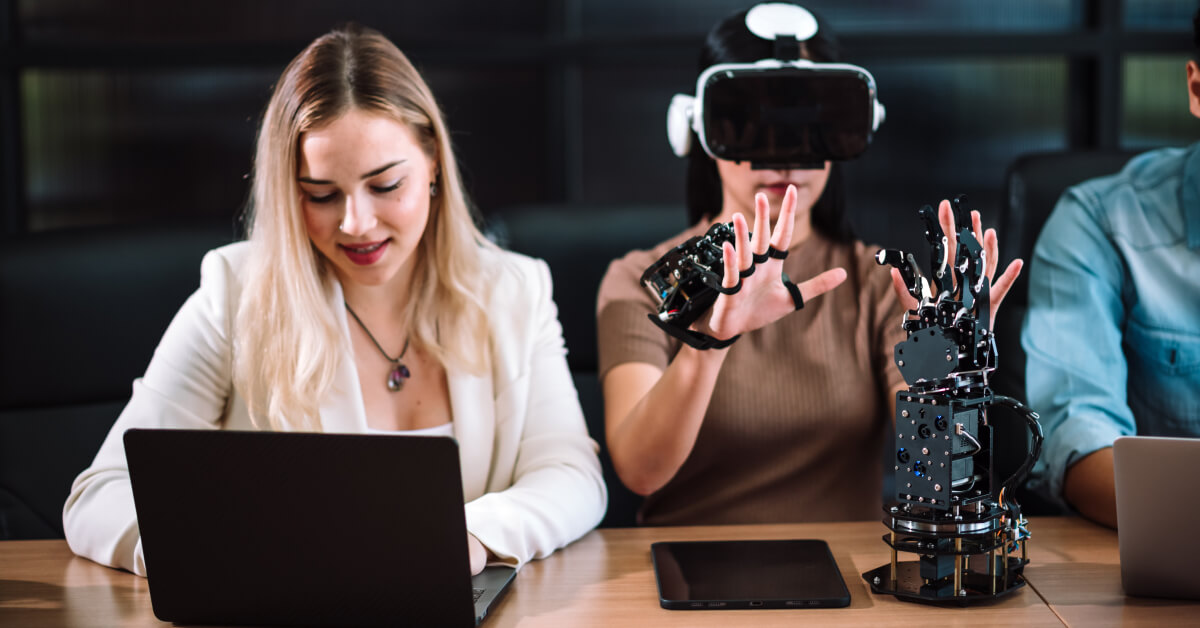 Woman using VR gloves and headset for robotics testing, while colleague works on laptop.