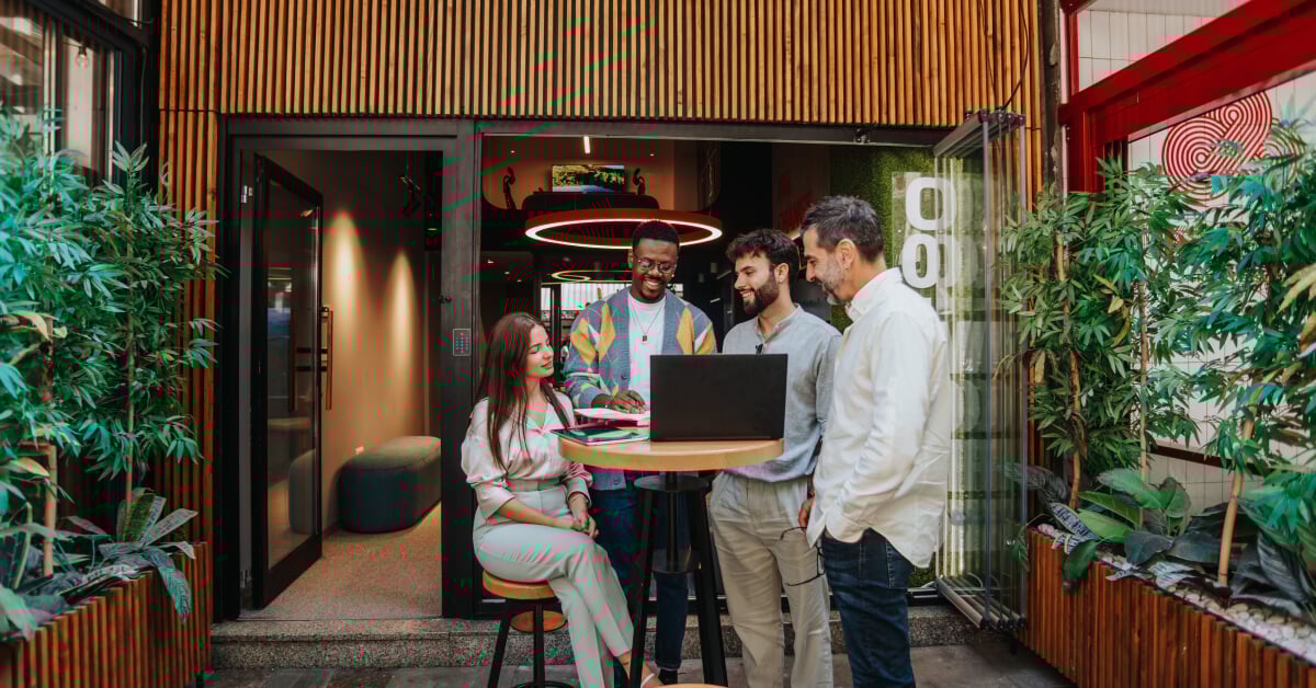 A diverse group of colleagues is gathered around a laptop during an outdoor meeting. The group consists of four men and one woman, all smiling and engaged in a collaborative discussion. They are standing and sitting around a high table in a modern outdoor space, with greenery and contemporary architecture in the background. The setting is casual and vibrant, reflecting a positive and creative work environment.