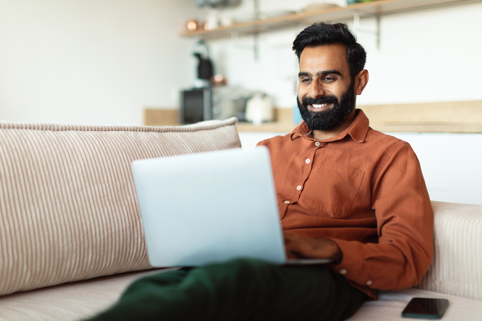 A man sitting on a couch smiles while shopping online using his laptop in a comfortable, modern home setting.