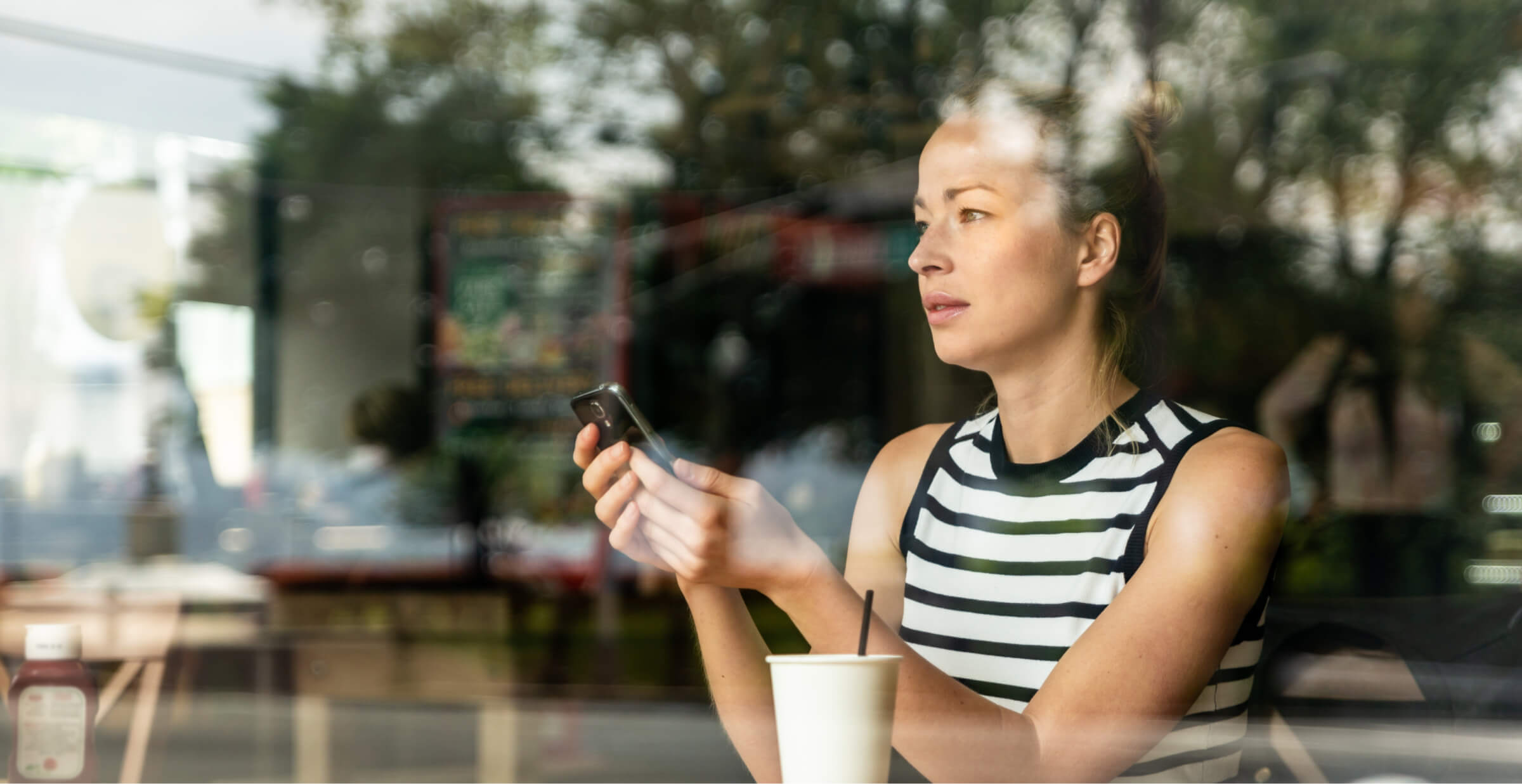 Woman using a smartphone in a cafe, seen through a glass window, with a contemplative expression.