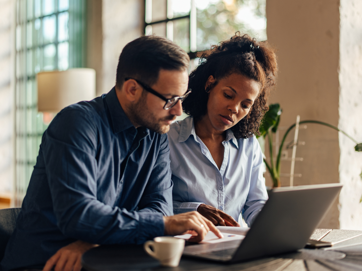 Two professionals collaborating at a table, reviewing documents on a laptop in a well-lit modern office space.