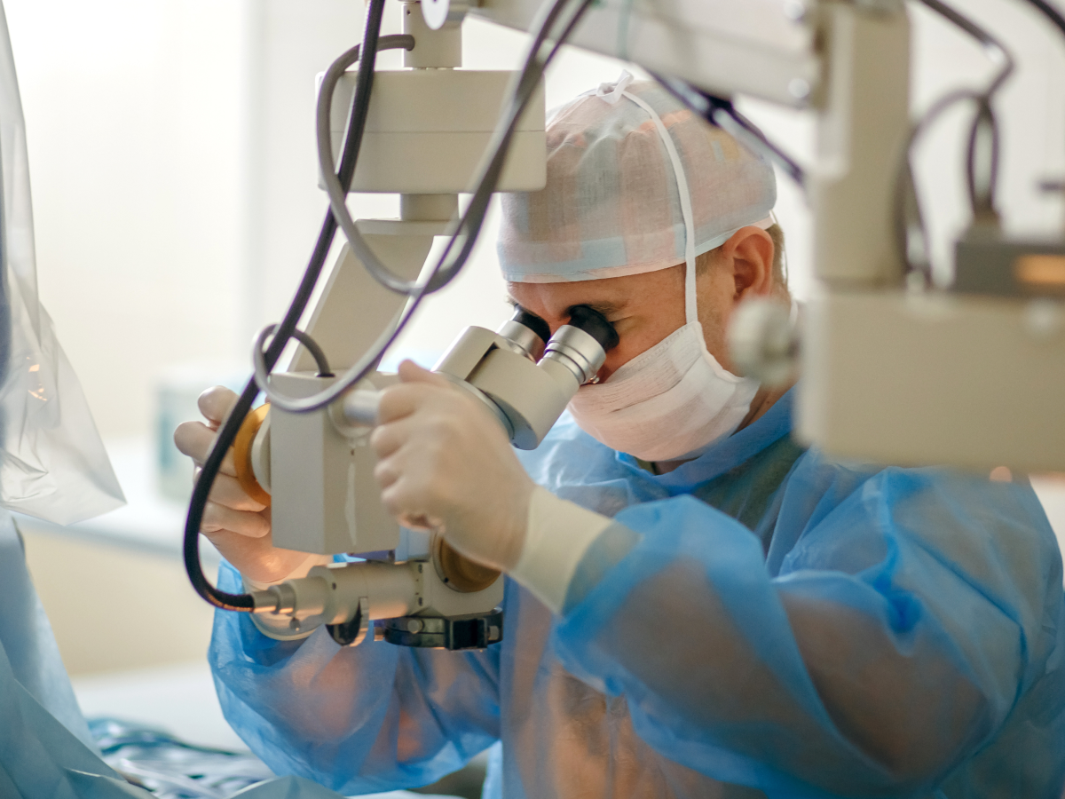A surgeon wearing a blue surgical gown, gloves, a face mask, and a hair cap is using a high-precision operating microscope during a medical procedure. The sterile environment and advanced equipment indicate a surgical setting, likely for ophthalmic or microsurgery.