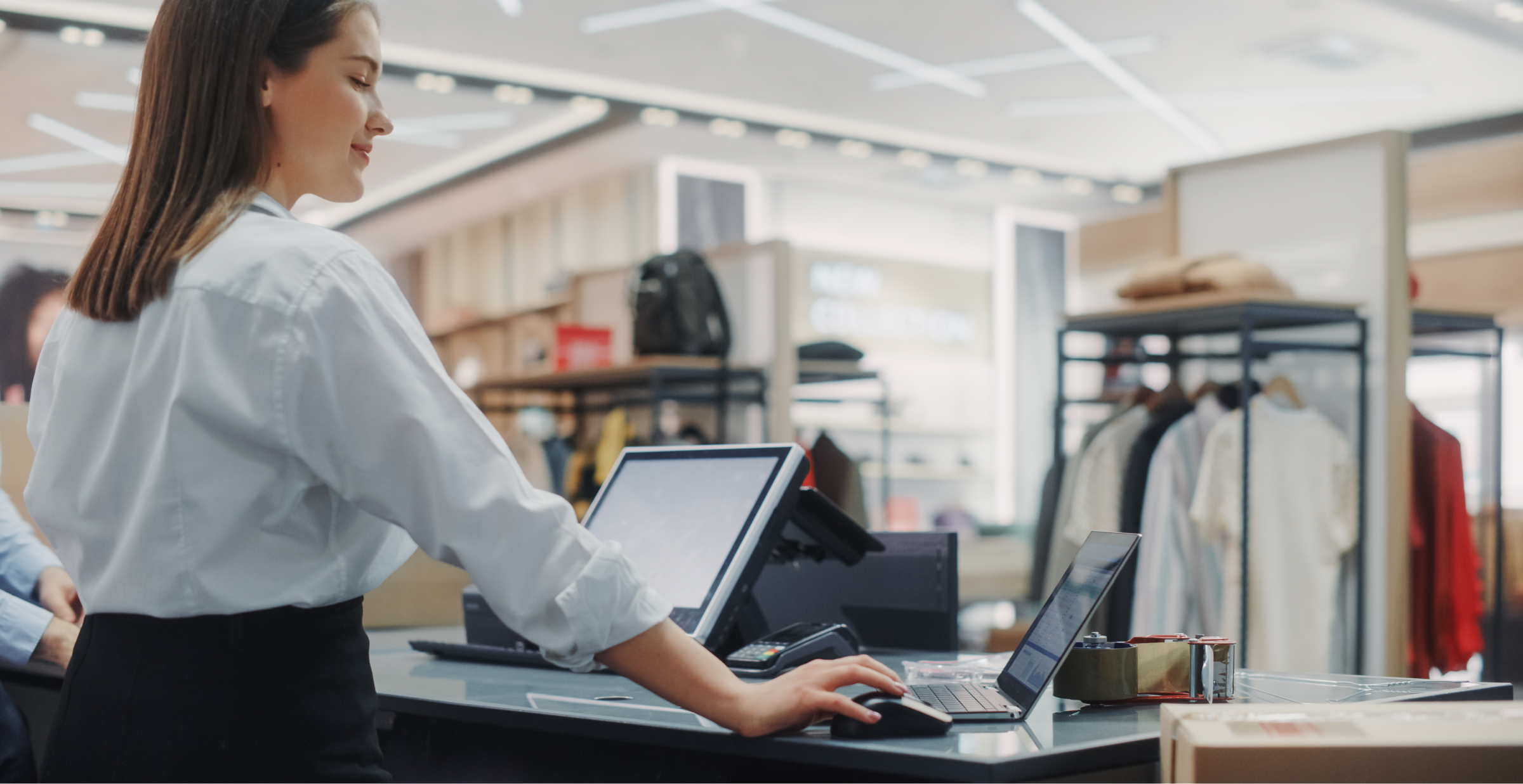  A retail store employee wearing a white shirt and black pants is working at a checkout counter. She is using a laptop and a touchscreen point-of-sale (POS) system. The background features clothing racks, shelves with bags, and a sign reading “NEW COLLECTION.” The store has a modern design with bright lighting and sleek fixtures.