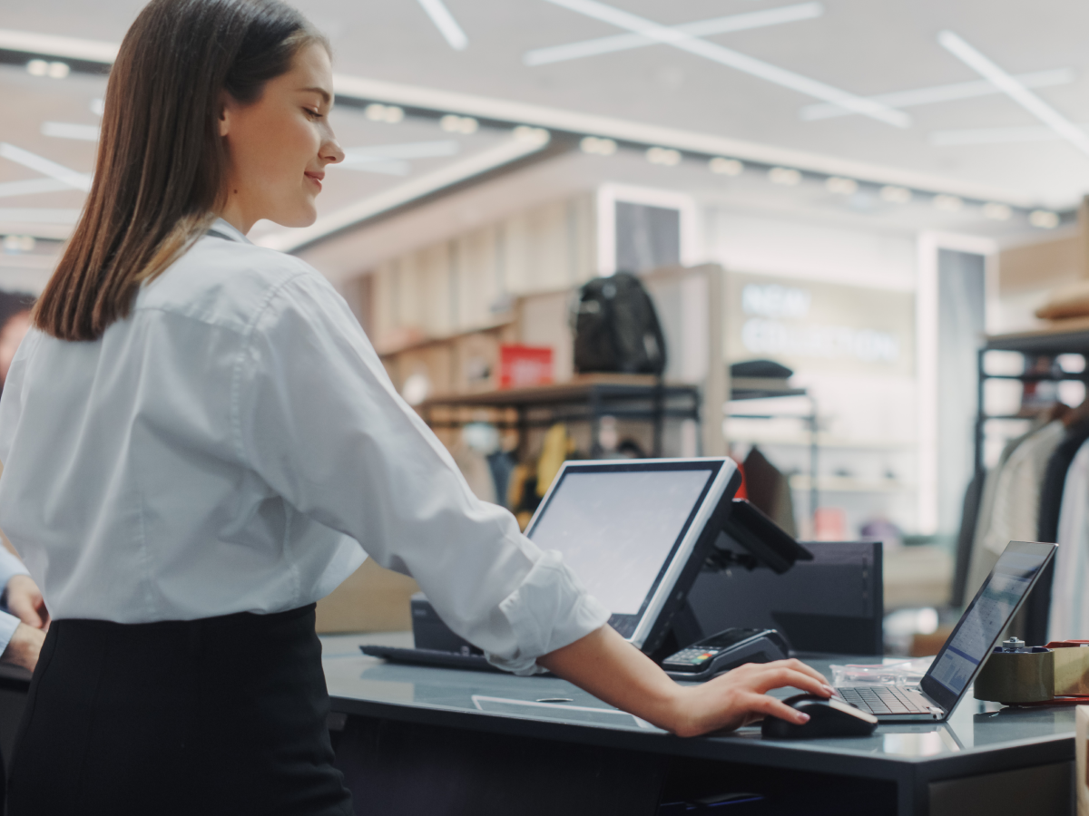  A retail store employee wearing a white shirt and black pants is working at a checkout counter. She is using a laptop and a touchscreen point-of-sale (POS) system. The background features clothing racks, shelves with bags, and a sign reading “NEW COLLECTION.” The store has a modern design with bright lighting and sleek fixtures.