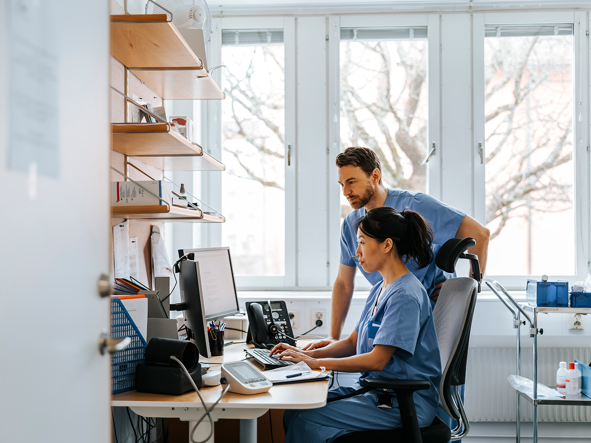 Two healthcare professionals in scrubs reviewing electronic health records on a computer in a clinical office setting.