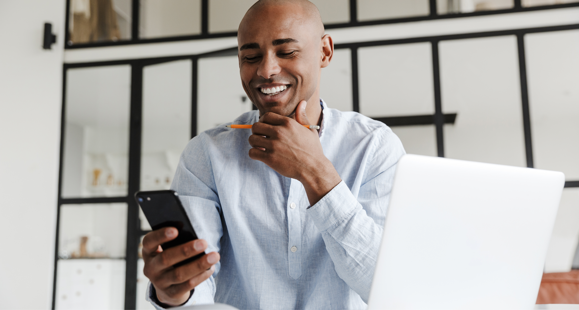 Smiling man in a light blue shirt holding a pencil and looking at his smartphone while seated at a desk with an open laptop in a modern office.
