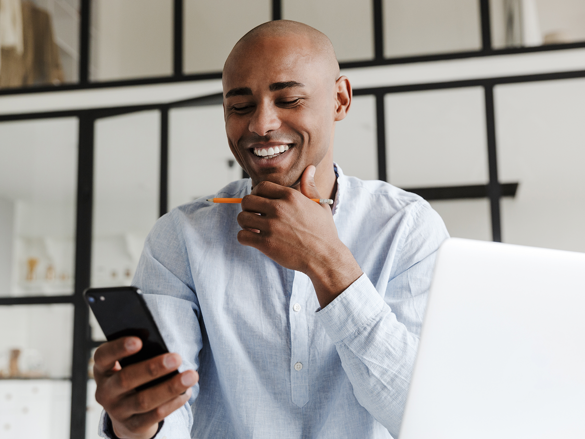 Smiling man in a light blue shirt holding a pencil and looking at his smartphone while seated at a desk with an open laptop in a modern office.