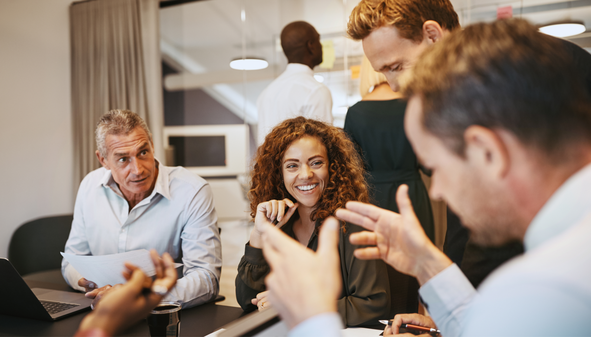 A dynamic office environment where a group of professionals engages in a lively discussion. A woman with curly hair and a bright smile is the center of attention, surrounded by colleagues who are actively contributing to the conversation. The setting features a modern workspace with natural lighting and a collaborative atmosphere. This image captures teamwork, engagement, and effective communication in a professional setting.
