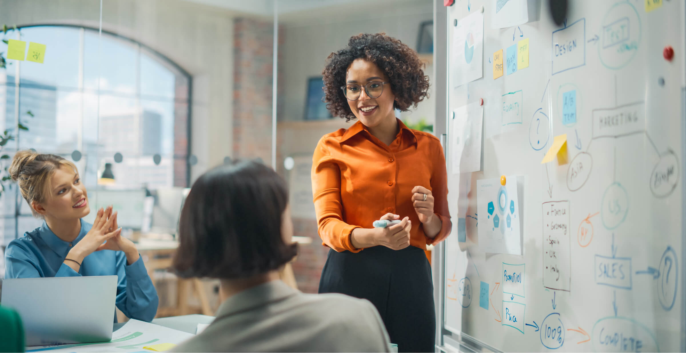 A businesswoman in an orange blouse leading a brainstorming session in a modern office, standing by a whiteboard covered with diagrams and sticky notes. Two colleagues are seated, engaged in the discussion, one of them smiling while listening attentively. The room has large windows and a bright, collaborative atmosphere.