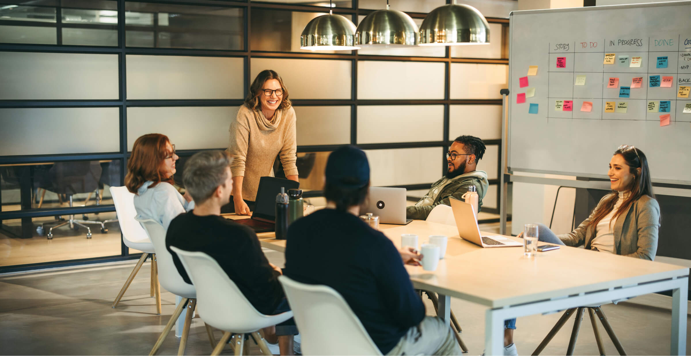 A group of colleagues sitting around a conference table, engaged in a collaborative meeting. A woman standing and smiling leads the discussion, while others listen and participate. The room has modern decor, with a whiteboard and laptops in use.