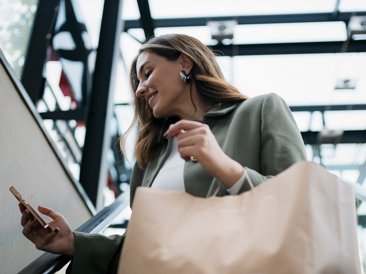 A woman holds a shopping bag while looking at her phone, appearing engaged in a mobile activity.  