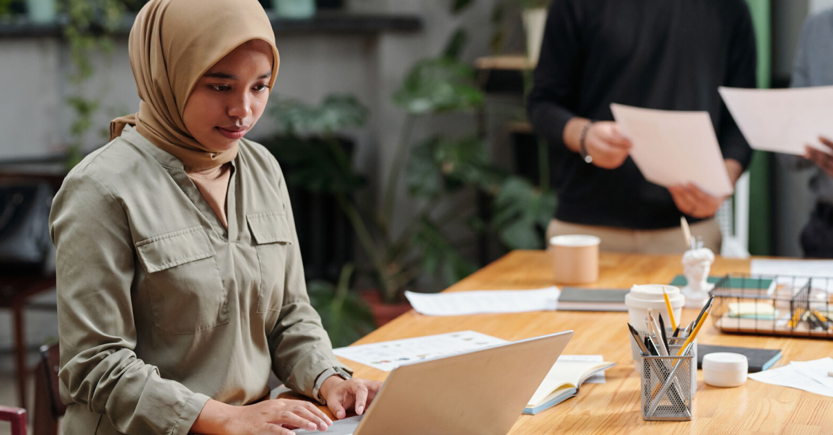 A young woman wearing a beige hijab and olive-green shirt is working on her laptop in a modern office space. In the background, colleagues are holding papers and discussing work at a wooden table filled with office supplies, coffee cups, and documents.