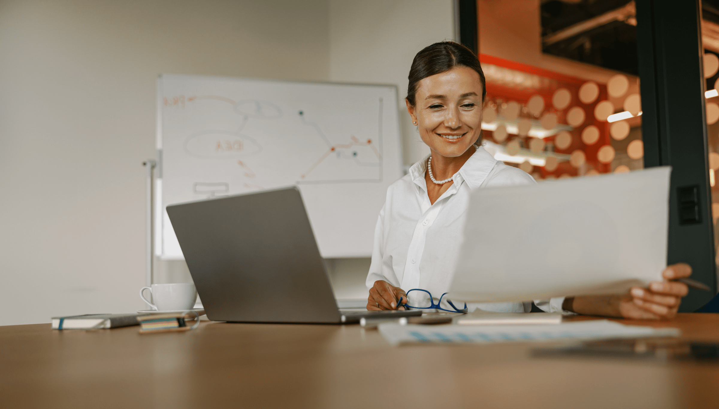 A woman is seated at a desk, smiling as she reviews a document in her hand. She has a laptop open in front of her, along with a cup of coffee, a notebook, and a pair of glasses resting on the table. Behind her, a whiteboard with diagrams is visible, and the background features a modern office setting with red and white polka-dot accents. The scene conveys a professional and positive work environment, with the woman appearing engaged and content with her work.