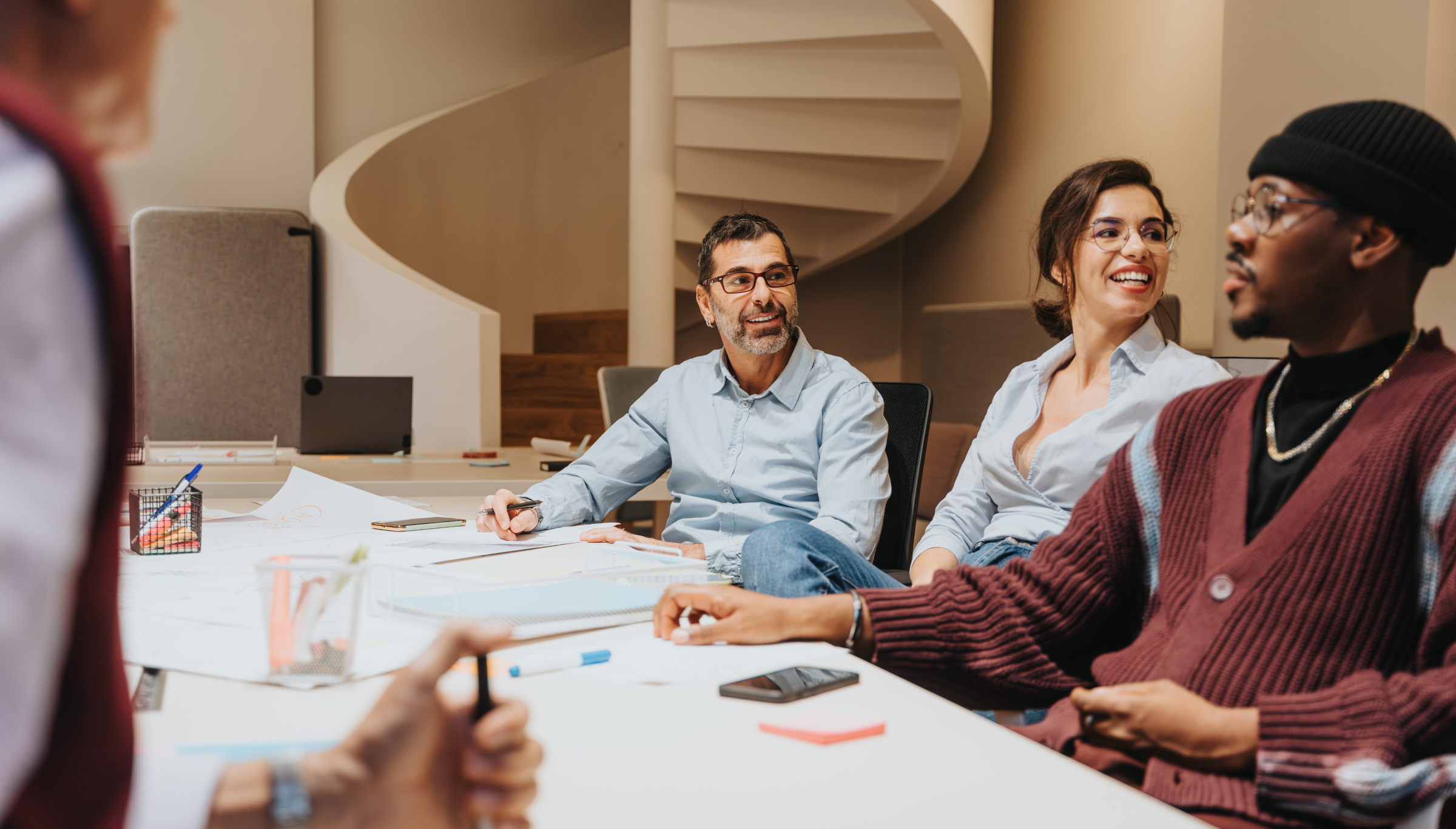 A group of people are having a lively discussion in a modern office setting. Three individuals, two men and one woman, are seated at a table, smiling and engaged in conversation. The table is covered with papers, pens, and a smartphone, suggesting a collaborative brainstorming session or meeting. The background features a spiral staircase, adding an architectural element to the workspace. The atmosphere is informal and creative, with the participants appearing relaxed and interactive.