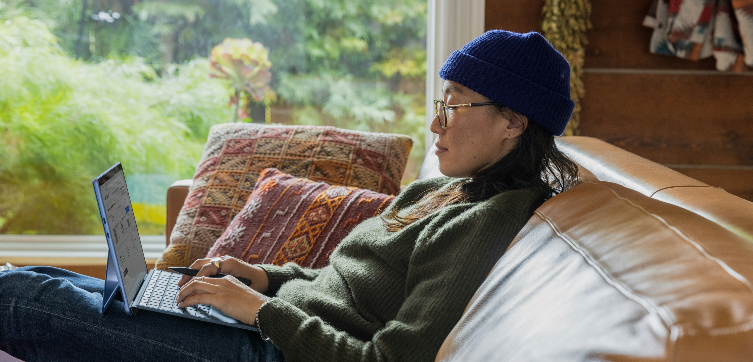 A person wearing glasses and a blue beanie is sitting comfortably on a leather couch, using a tablet. The scene is cozy, with colorful cushions behind them and a large window offering a view of lush greenery outside. The person appears relaxed and focused on their work or browsing, suggesting a peaceful, home-based work or leisure environment.