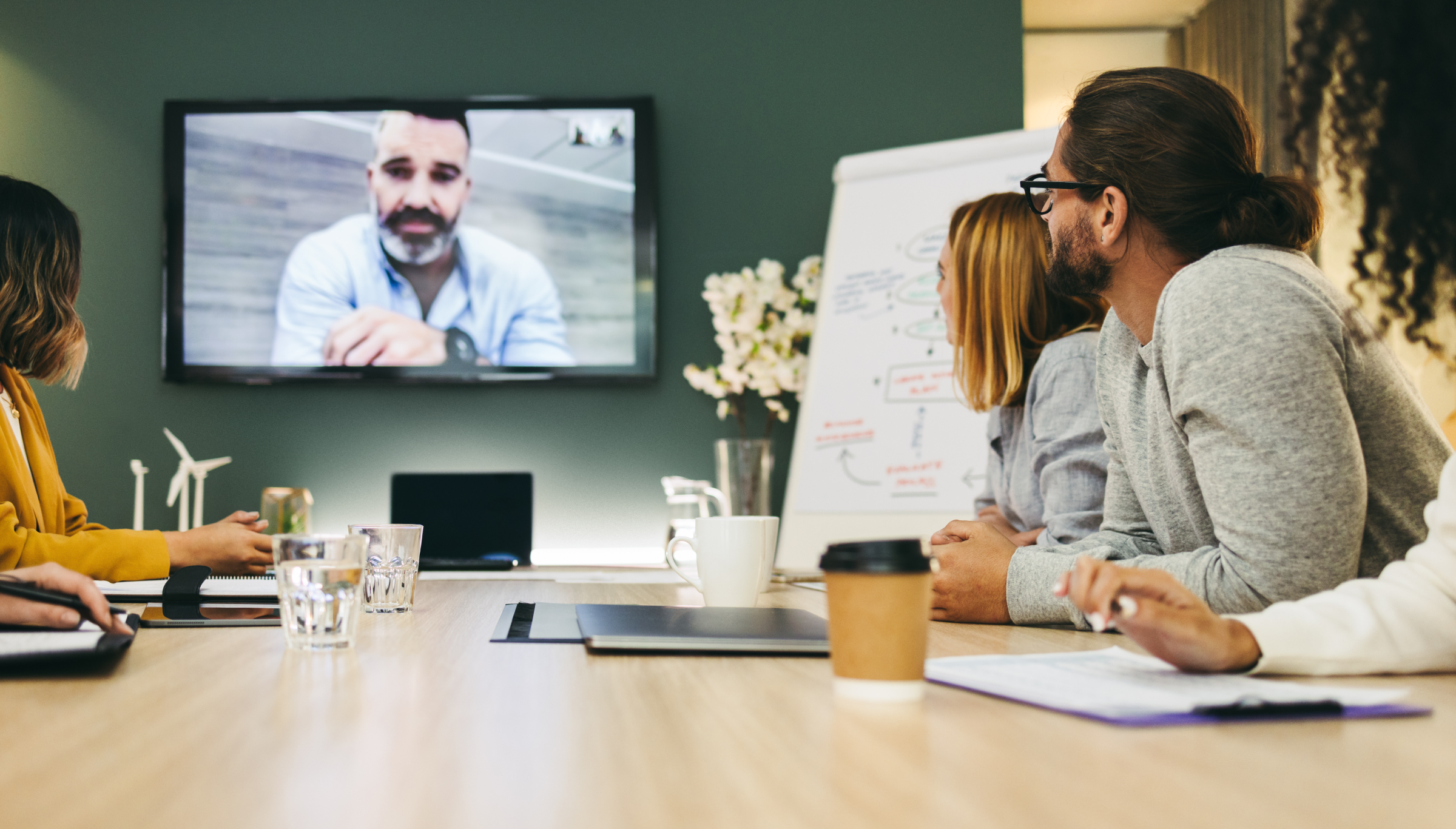 A group of people are seated around a conference table in a modern meeting room, participating in a video conference. A large screen on the wall displays a man speaking during the video call. The team members are attentively watching and taking notes, with a whiteboard full of diagrams and notes visible in the background. The table is equipped with laptops, notebooks, coffee cups, and water glasses, indicating an engaged and collaborative work session. The atmosphere is professional and focused, blending in-person and virtual communication.