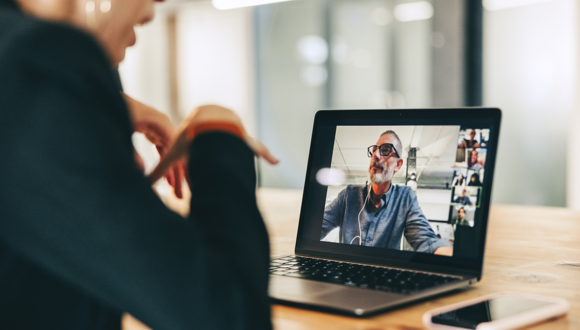 A person is participating in a video call on a laptop, with the screen showing multiple participants in a virtual meeting. The primary focus on the screen is a man wearing glasses and using earphones, speaking during the call. The laptop is placed on a wooden table, with a smartphone nearby. The setting appears to be a casual workspace, emphasizing remote collaboration and communication in a professional environment. The person on the video call is actively engaged in the discussion.