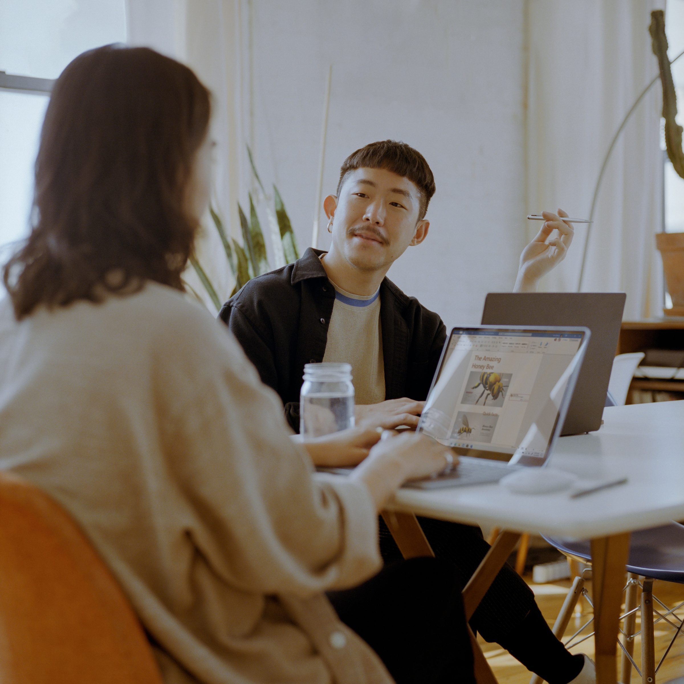 Two people are collaborating at a table in a bright, modern workspace. One person, holding a stylus, is looking at the other while discussing their work. The second person is focused on their laptop, which displays an article about bees. A mason jar is on the table, adding a casual touch to the setting. The atmosphere is creative and relaxed, with natural light filling the room and plants in the background, suggesting a comfortable and inspiring environment for collaboration.