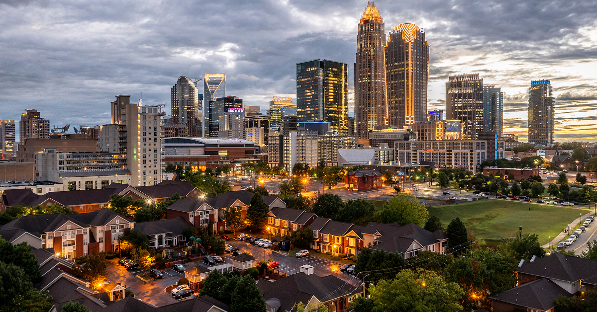 Evening view of Charlotte, North Carolina skyline with illuminated buildings and nearby residential neighborhood, highlighting urban tourism appeal.