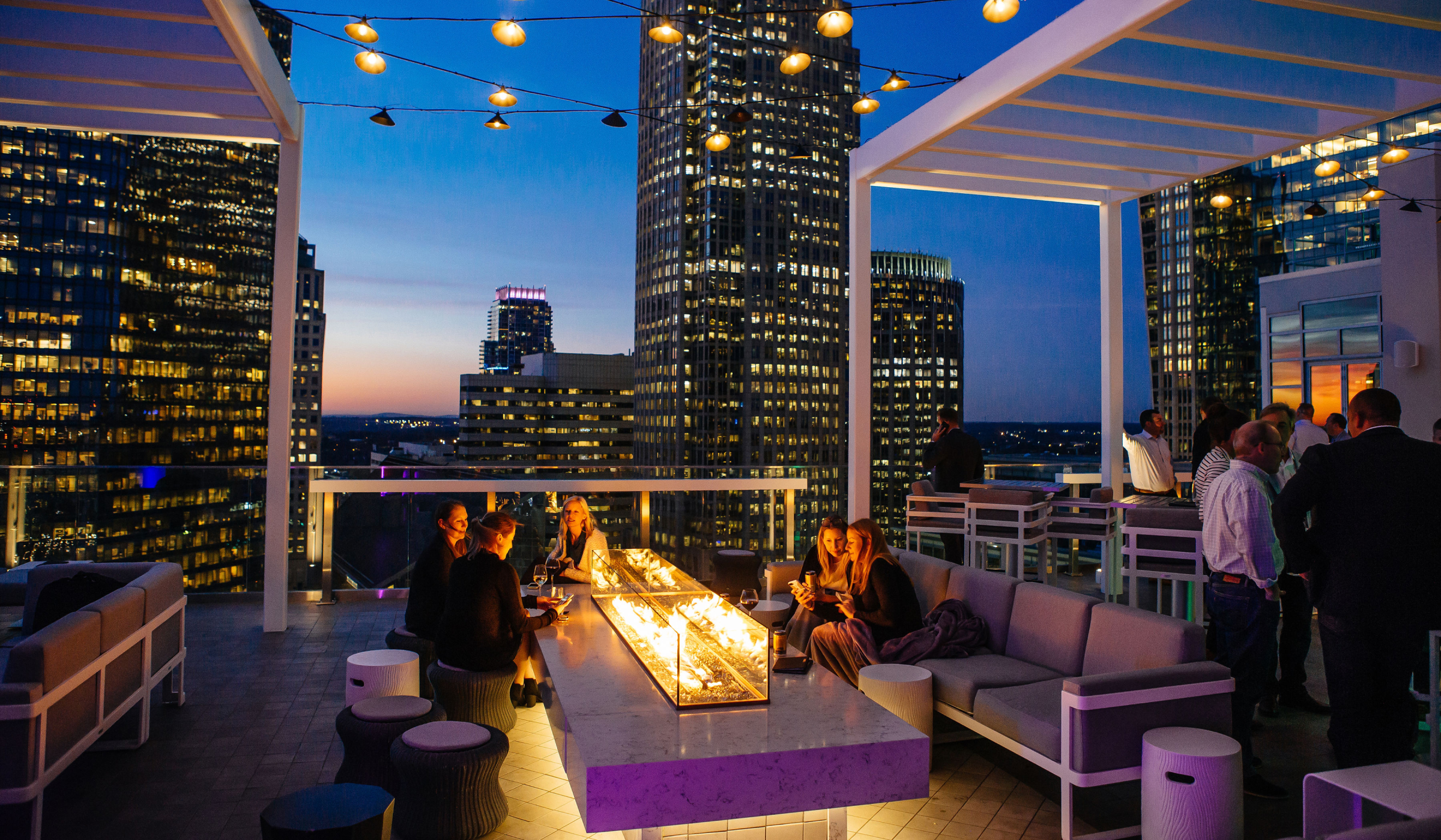 People socializing at a rooftop bar in Charlotte, NC at sunset, with city skyscrapers lit up in the background and a modern fire pit centerpiece.