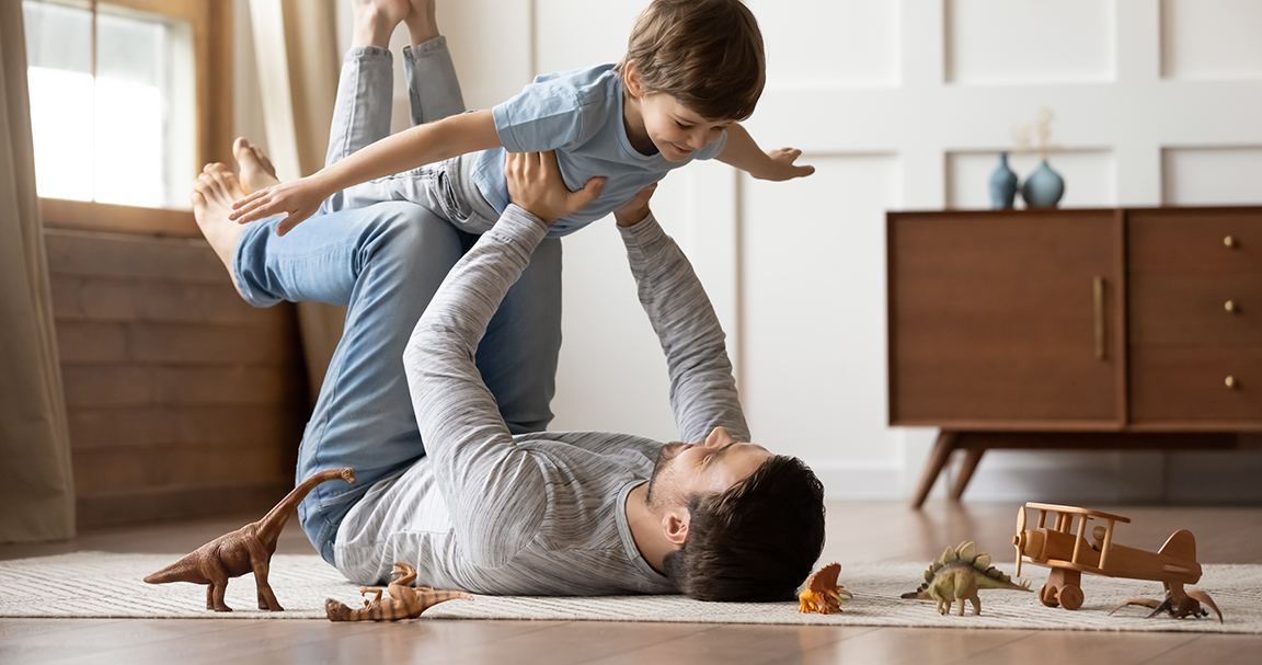 Caucasian man lifts young child in child's bedroom surrounded by toy dinosaurs and wooden airplane