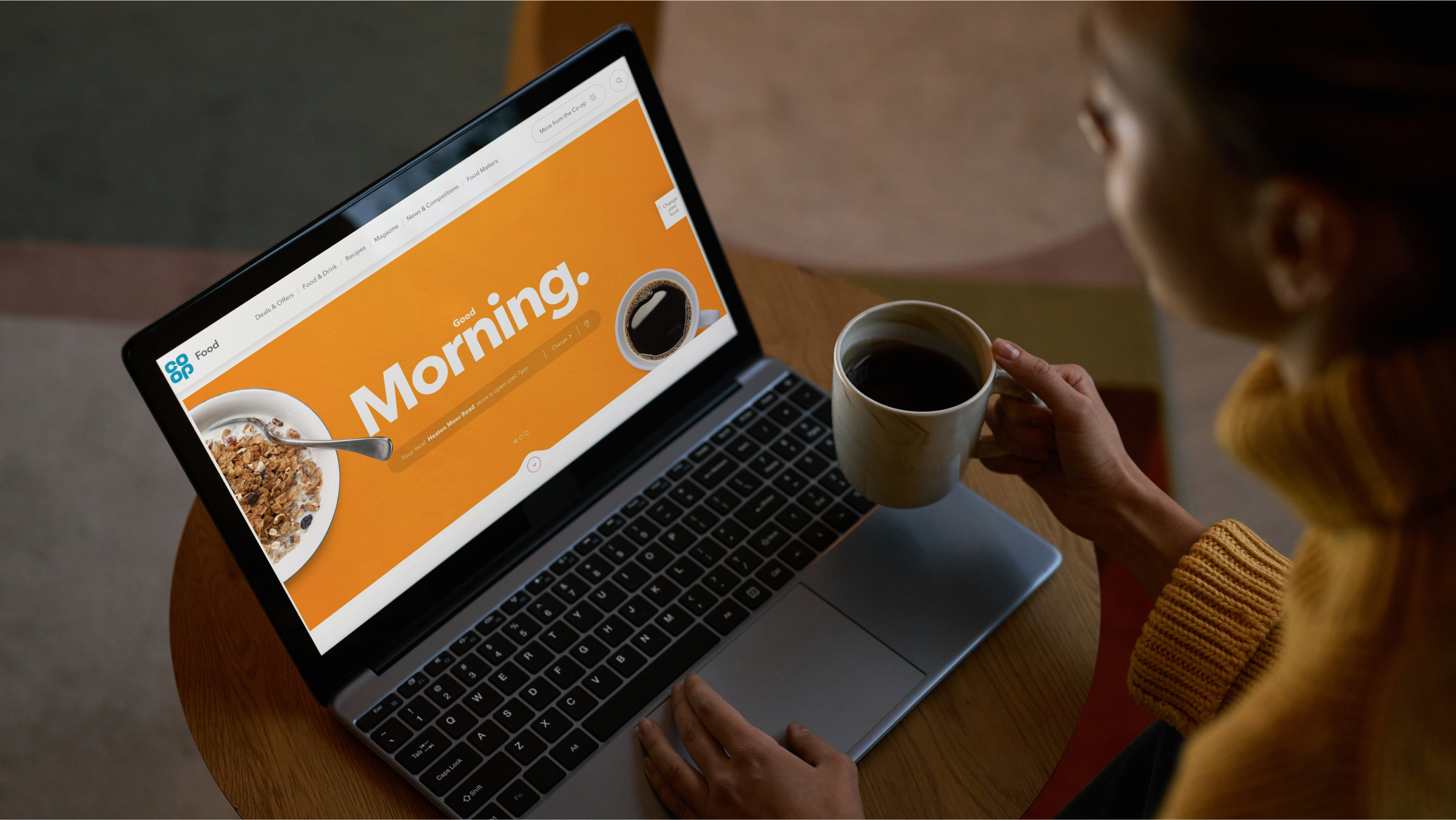 A person sitting at a table, holding a cup of coffee while browsing a website on a laptop. The laptop screen displays a bright orange background with the text 'Good Morning,' along with images of a coffee cup and a bowl of cereal. The setting suggests a relaxed morning routine.