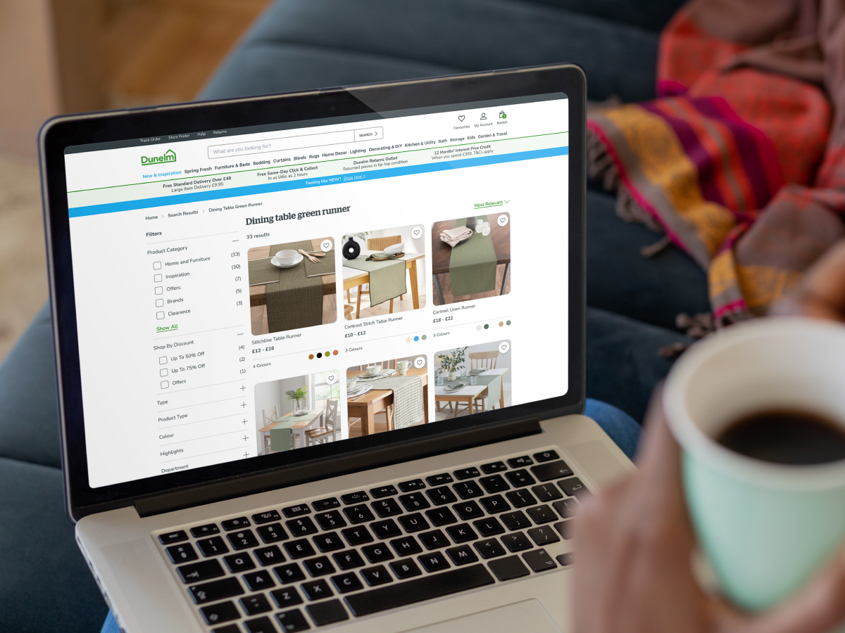 A person holding a cup of coffee while browsing an online store on a laptop. The screen displays a Dunelm website page showing various green dining table runners with product images, prices, and filters for selection. The individual is seated on a couch with a colorful throw blanket in the background.