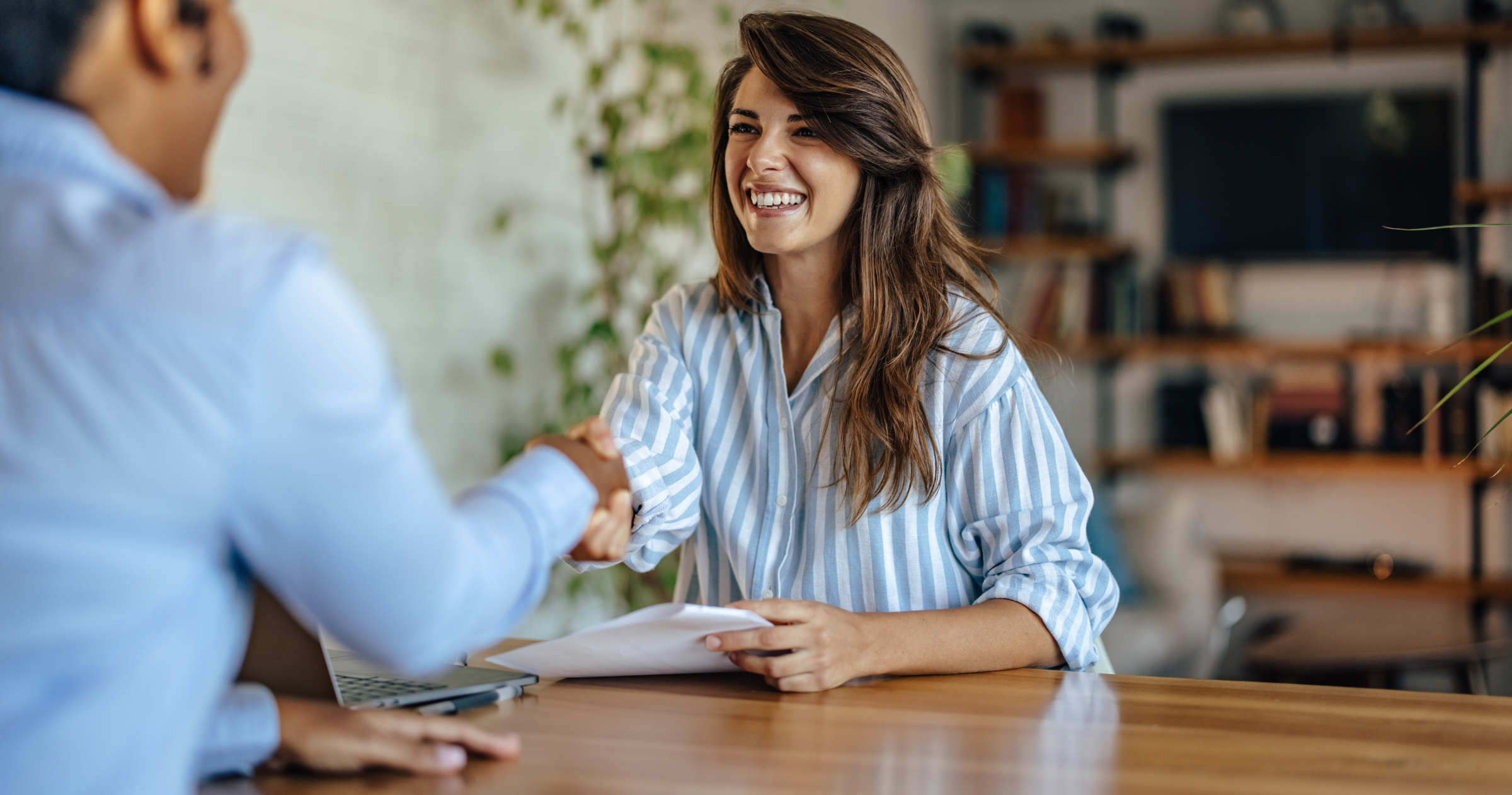 A smiling woman in a striped shirt reaching across a desk to shake hands with a man barely in the frame.