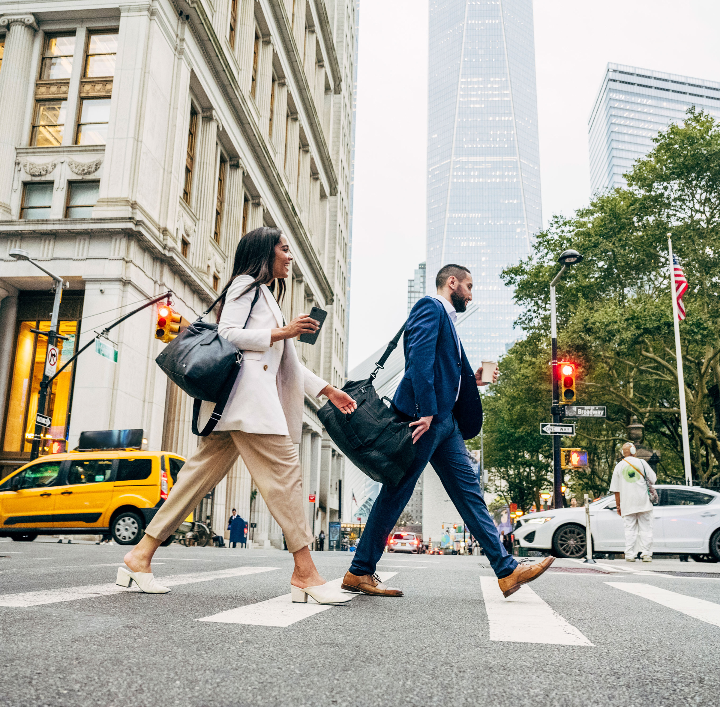 Two pedestrians crossing a busy street in New York City, surrounded by tall buildings and city traffic.  