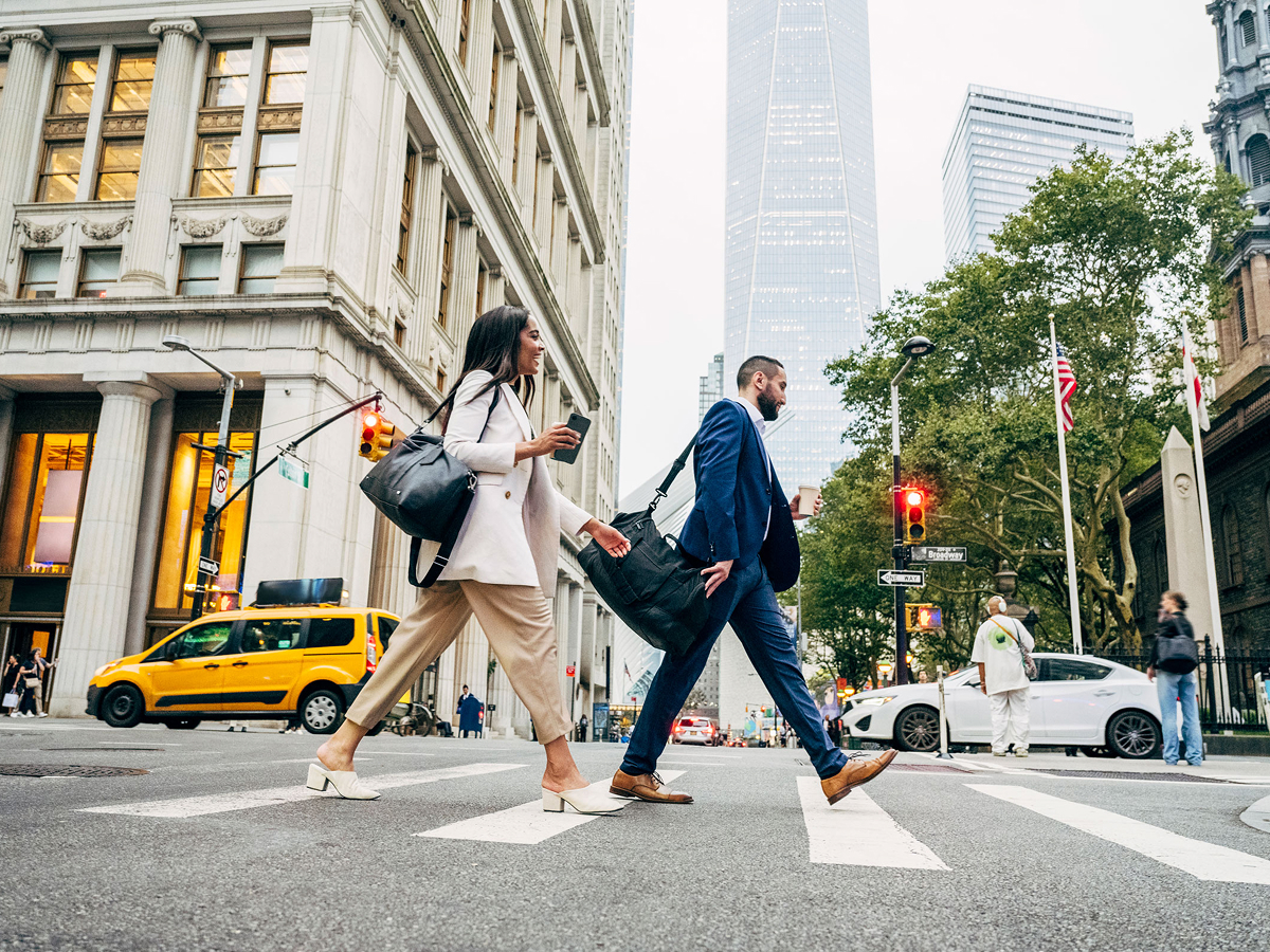Two pedestrians crossing a busy street in New York City, surrounded by tall buildings and city traffic.  