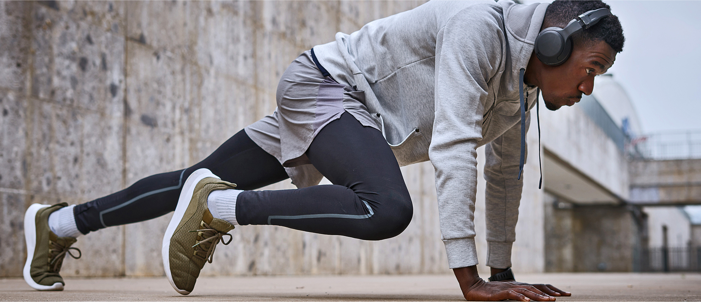 A focused male athlete wearing a hoodie and headphones is stretching outdoors. He is in a dynamic stretching position, preparing for a workout or run, with one leg forward and the other extended back, both hands on the ground. The background features an urban setting with concrete walls, suggesting a city workout environment. The image captures the athlete’s dedication and readiness for physical activity.