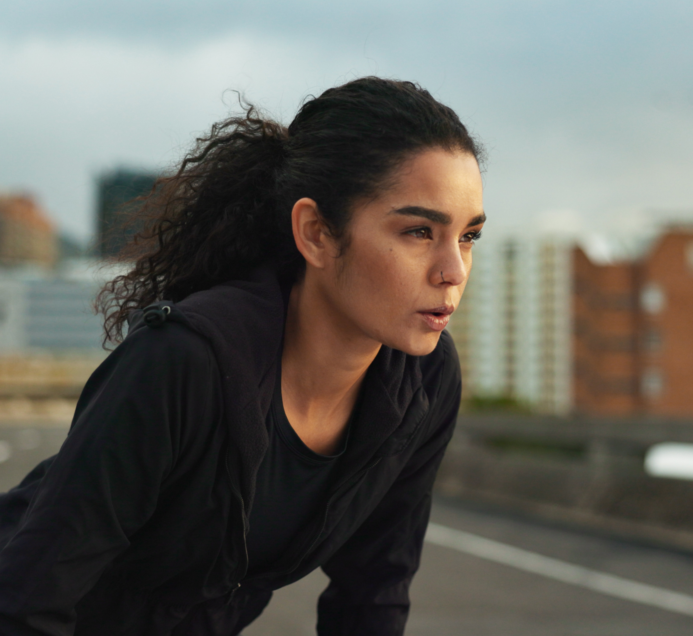 A determined female runner in athletic gear is taking a break after a run. She leans forward, catching her breath, with a focused expression on her face. The background shows an urban setting with blurred buildings, suggesting an early morning or late afternoon workout. The image captures her dedication and perseverance, embodying the spirit of fitness and resilience.