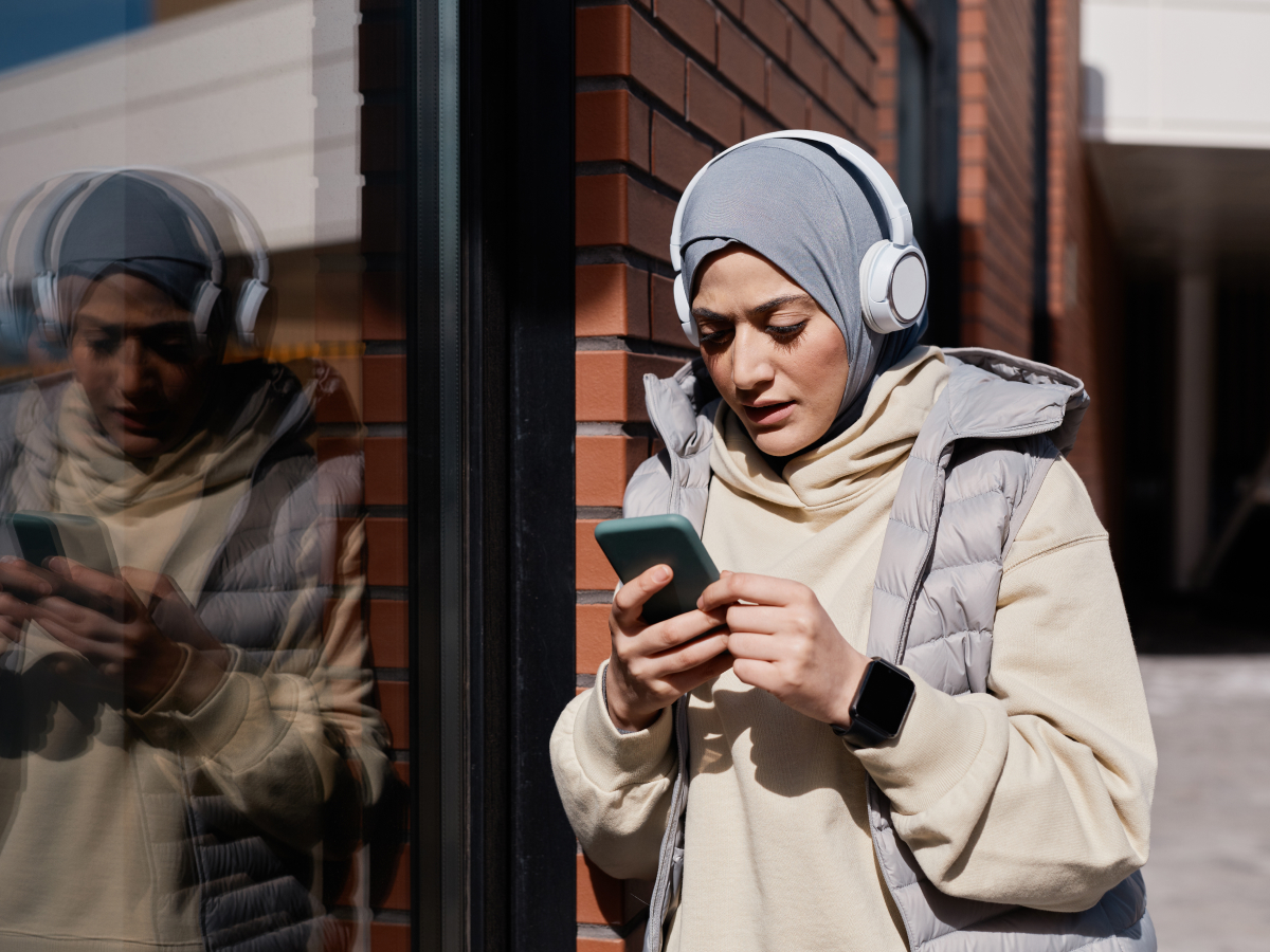 A woman wearing a hijab, headphones, and a puffy vest, standing against a brick wall outdoors, looking at her smartphone while her reflection appears on the glass next to her.