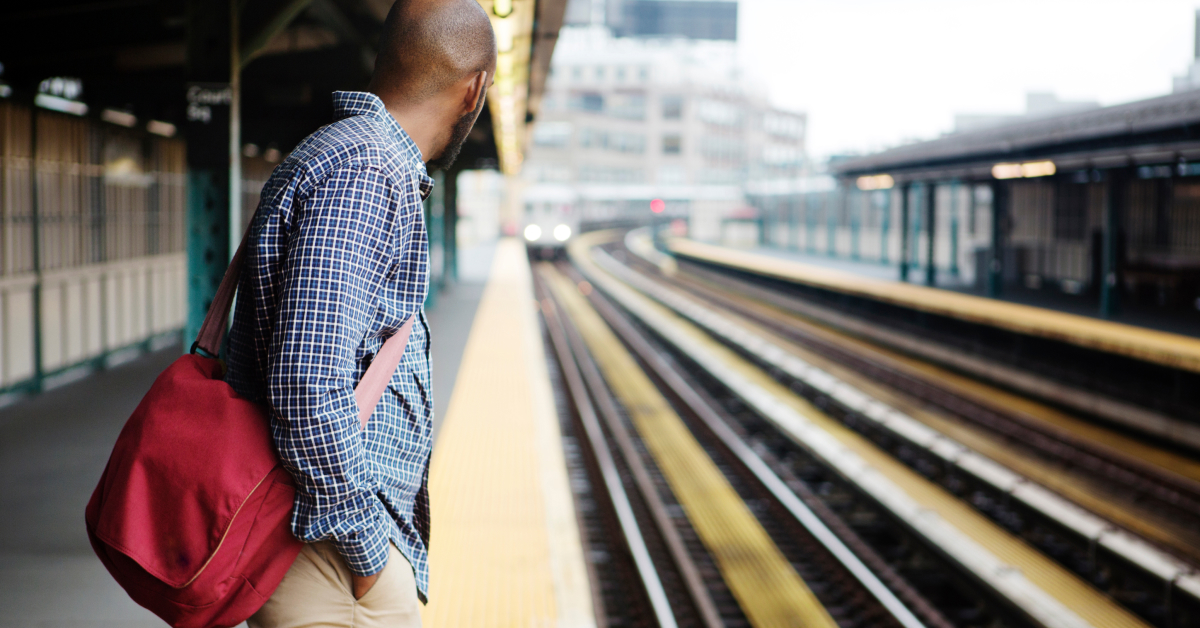 A man wearing a checkered shirt and carrying a red bag stands on a train platform, looking down the tracks as a train approaches in the distance.