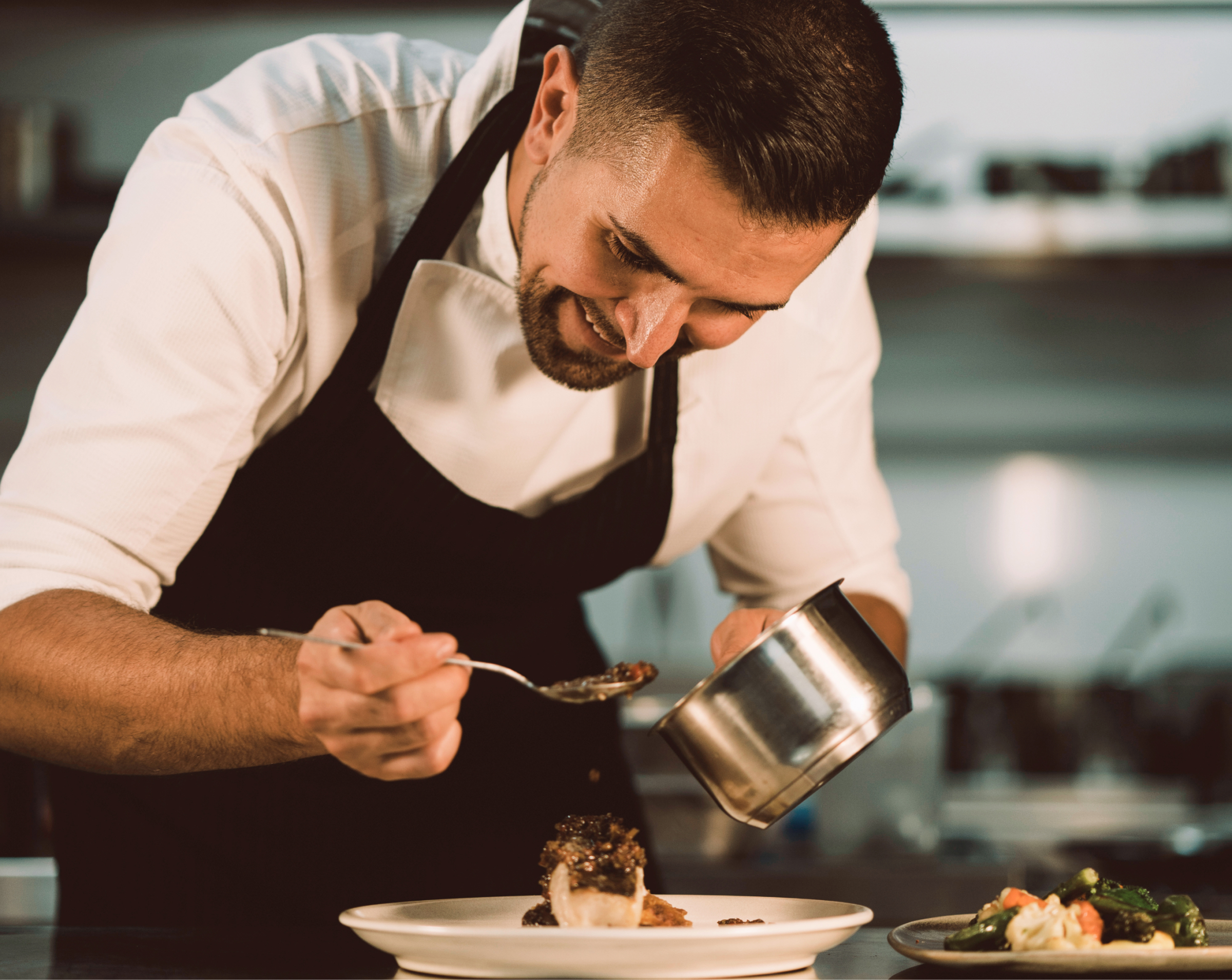 A professional chef is smiling as he carefully plates food in a kitchen. He is wearing a white chef’s coat and a black apron, using a spoon to delicately arrange the dish. The scene captures the chef’s attention to detail and passion for his craft, highlighting the artistry and precision involved in creating a culinary masterpiece. The background suggests a professional kitchen environment, emphasizing the chef’s expertise.