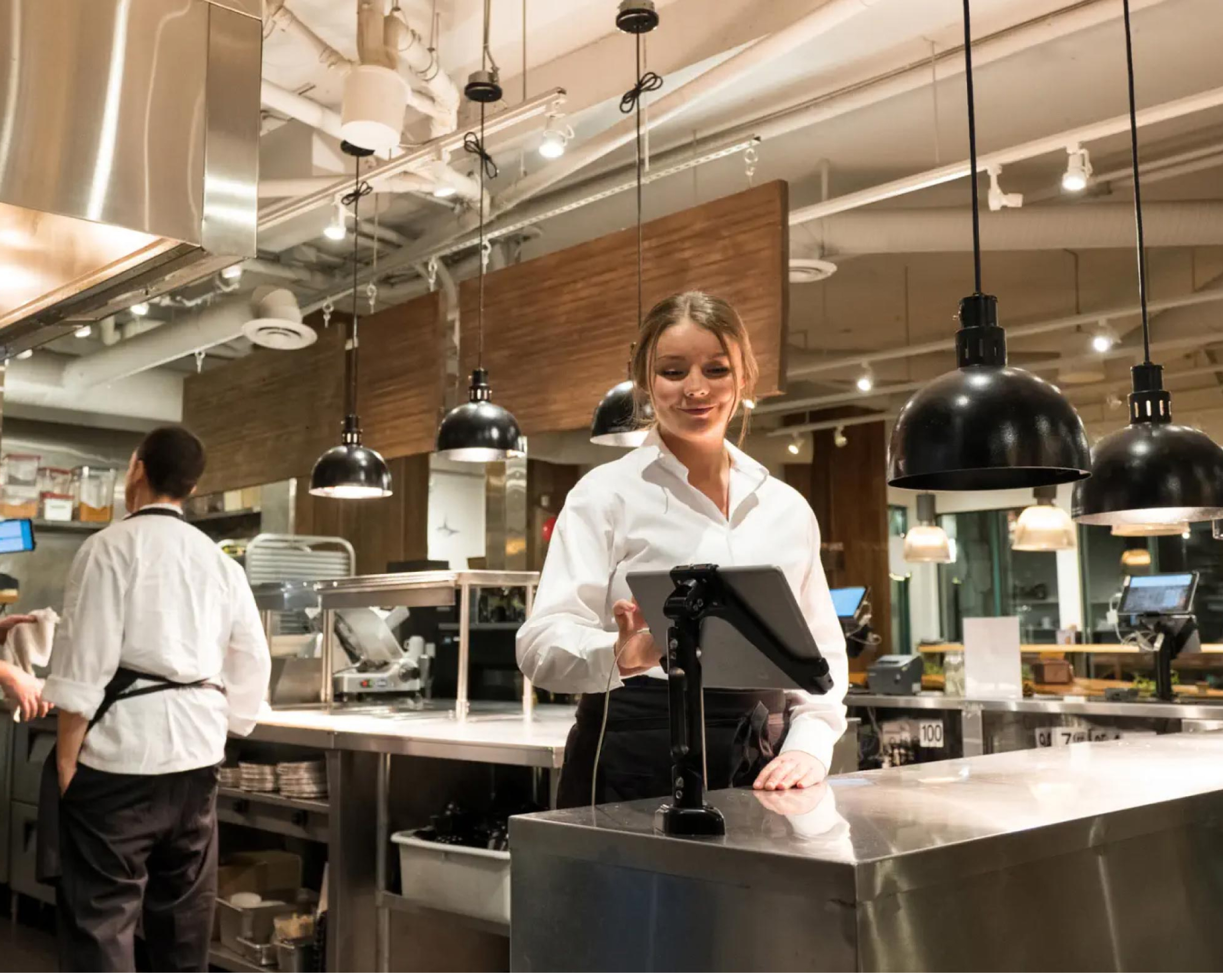 A server in a white shirt standing in a modern, well-lit kitchen, smiling as she interacts with a mounted tablet. The kitchen features industrial-style lighting and stainless steel surfaces, with another staff member working in the background.