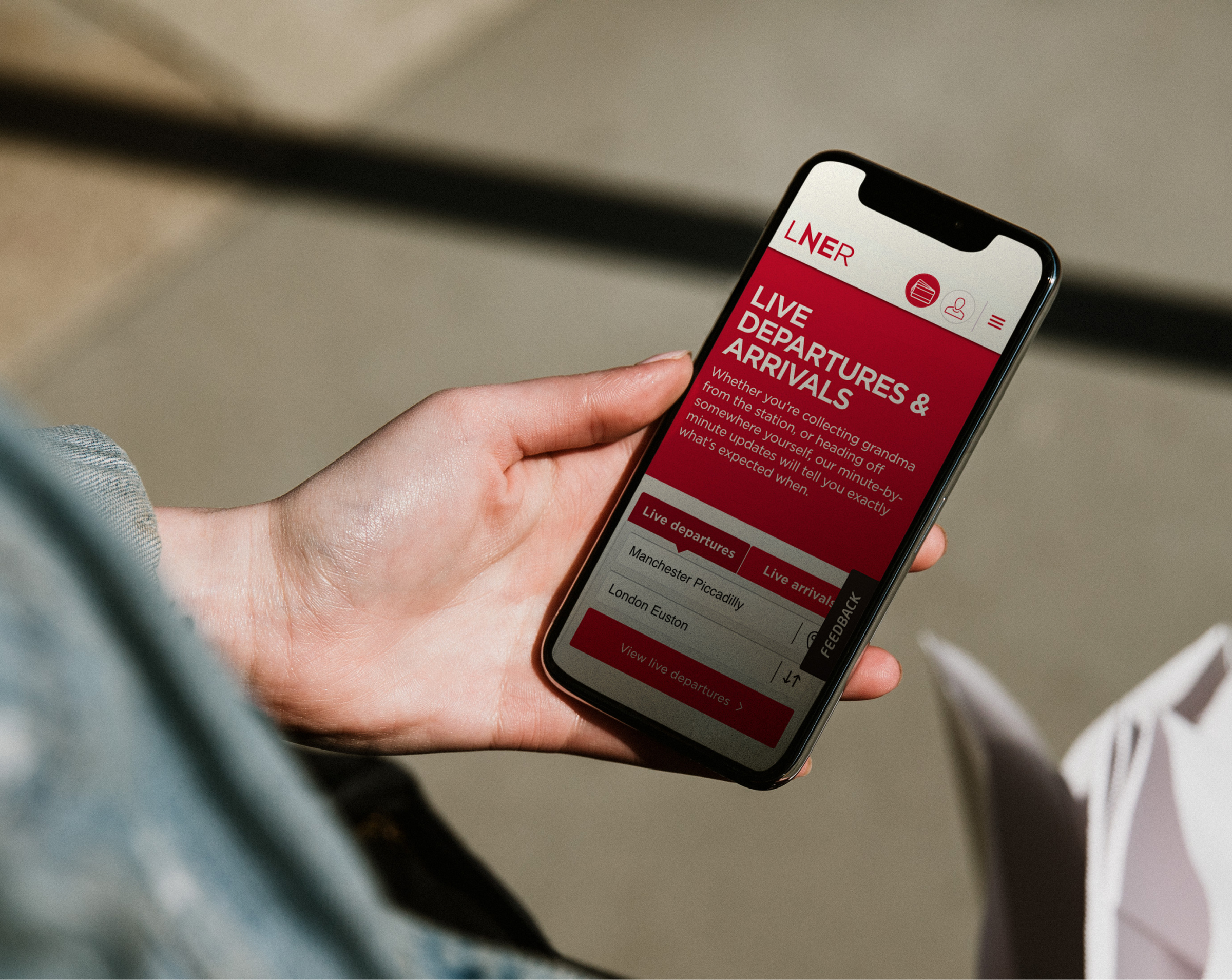 A person holding a smartphone displaying the LNER app, which shows live departures and arrivals information for train stations, with a focus on Manchester Piccadilly and London Euston.