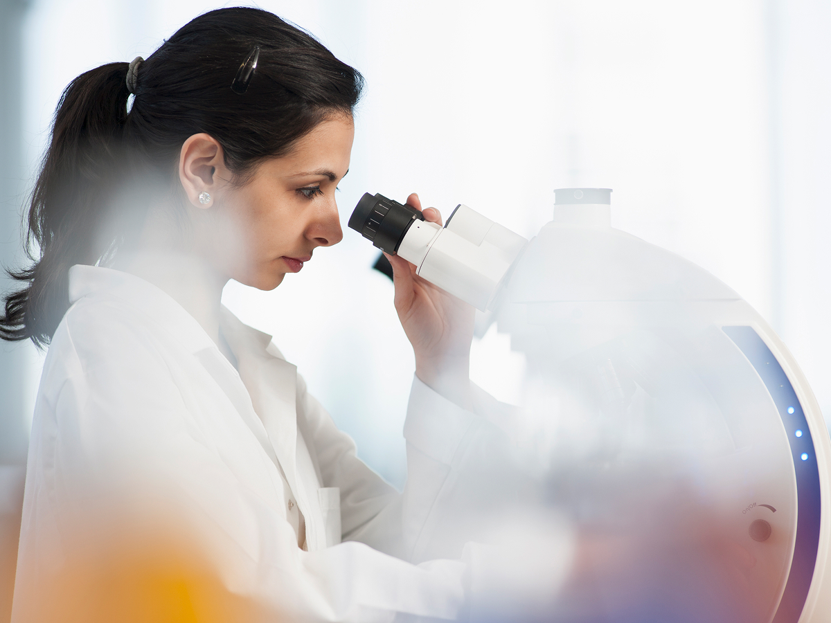 A woman in a white lab coat peers into a white microscope in a bright laboratory, with her dark hair pulled back and her focus on the eyepiece as soft light filters through a window.