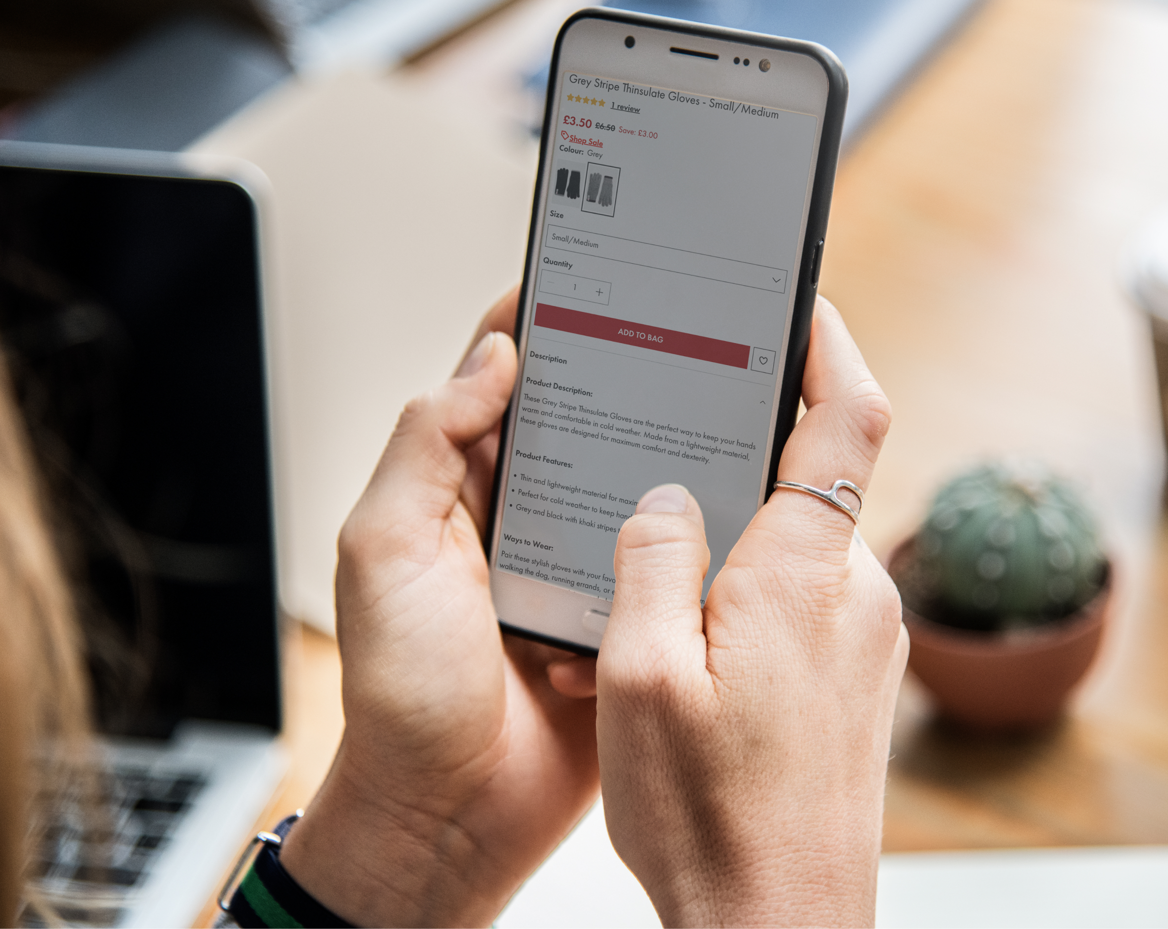 A person holding a smartphone, viewing a product page for Grey Stripe Thinsulate Gloves. The screen shows the product description, price, and purchasing options. In the background, a laptop and a small potted cactus are visible, suggesting a casual or work-from-home environment.