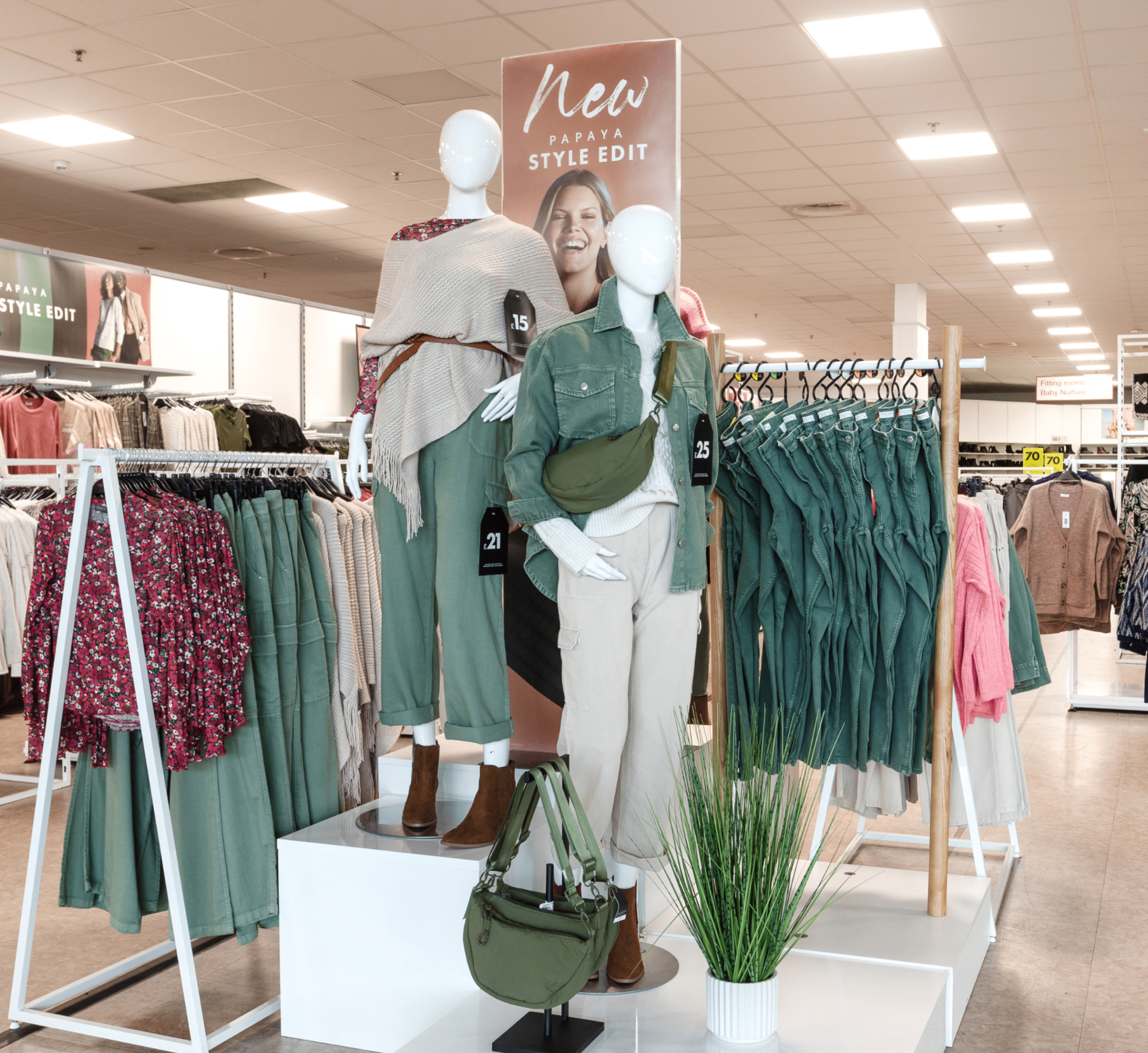 A display inside a retail store featuring the 'New Papaya Style Edit' collection. Mannequins are dressed in neutral and green tones, showcasing casual outfits with accessories like a green crossbody bag. Racks of matching clothing surround the display, with a modern, stylish vibe.