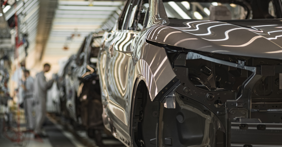 An automotive car assembly line in a factory with several vehicles in various stages of construction and workers blurred in the background.