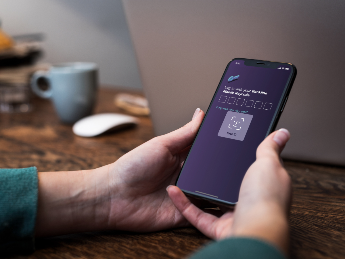 A person holding an iPhone displaying a log in screen. The FaceID functionality of iOS is displayed, together with a heading that reads “Log in with your Bankline Mobile Keycode”, followed by six empty number fields. Below there’s a text link saying “Forgotten your Keycode?”. 