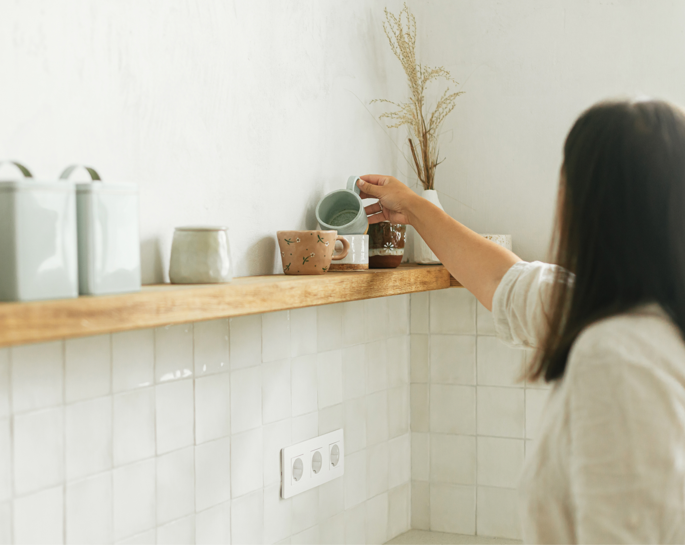 A person is seen from behind while placing a light blue ceramic cup on a wooden shelf in a minimalistic kitchen. The shelf is decorated with various ceramic mugs, jars, and a vase with dried plants. The walls have a light tone, and the tiled backsplash is white. The overall decor exudes a clean, modern, and cozy aesthetic.