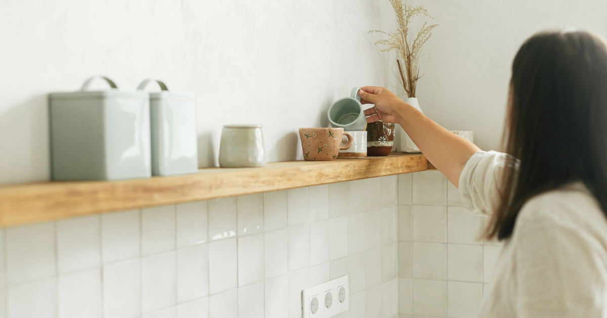 A person is seen from behind while placing a light blue ceramic cup on a wooden shelf in a minimalistic kitchen. The shelf is decorated with various ceramic mugs, jars, and a vase with dried plants. The walls have a light tone, and the tiled backsplash is white. The overall decor exudes a clean, modern, and cozy aesthetic.