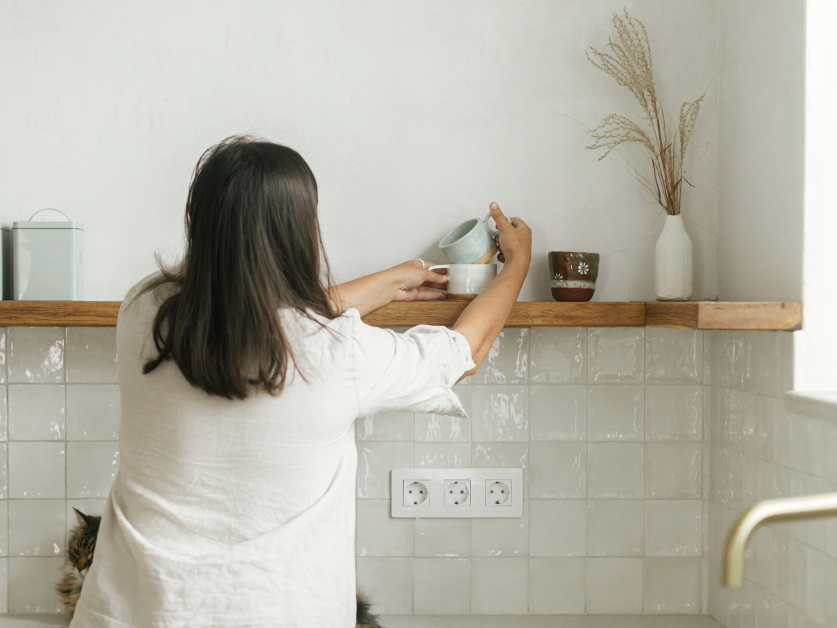 A person is seen from behind while placing a light blue ceramic cup on a wooden shelf in a minimalistic kitchen. The shelf is decorated with various ceramic mugs, jars, and a vase with dried plants. The walls have a light tone, and the tiled backsplash is white. The overall decor exudes a clean, modern, and cozy aesthetic.